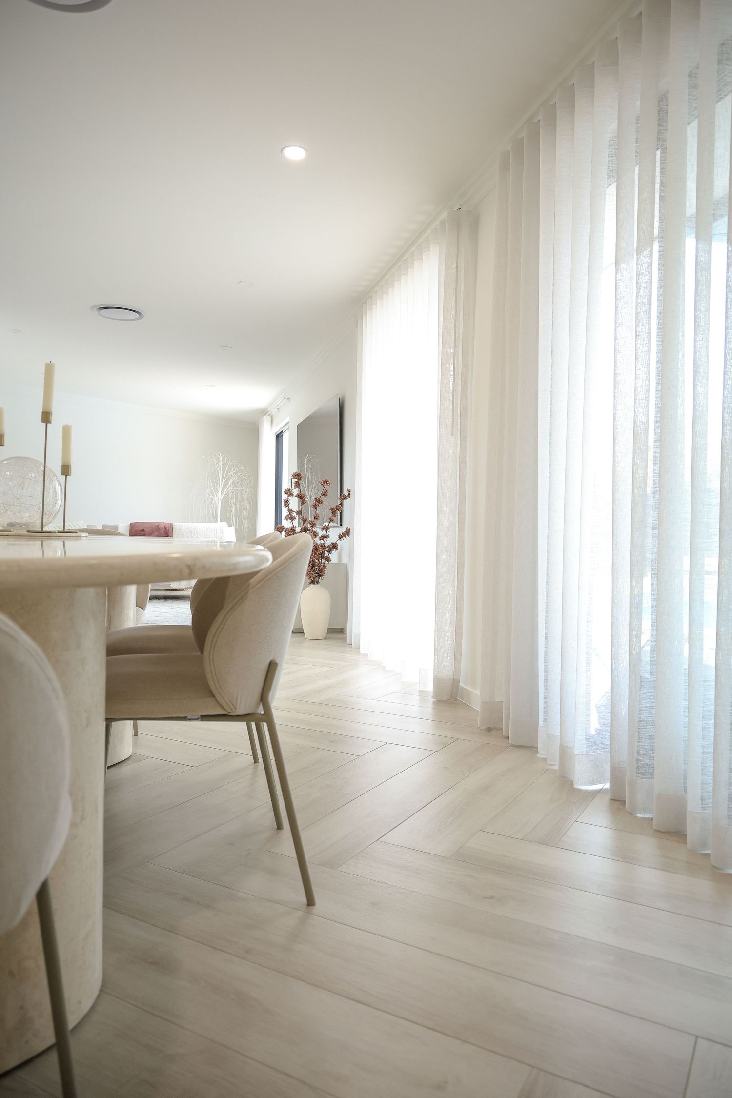 Modern dining room with light wood flooring, white chairs, and sheer curtains letting in natural light.