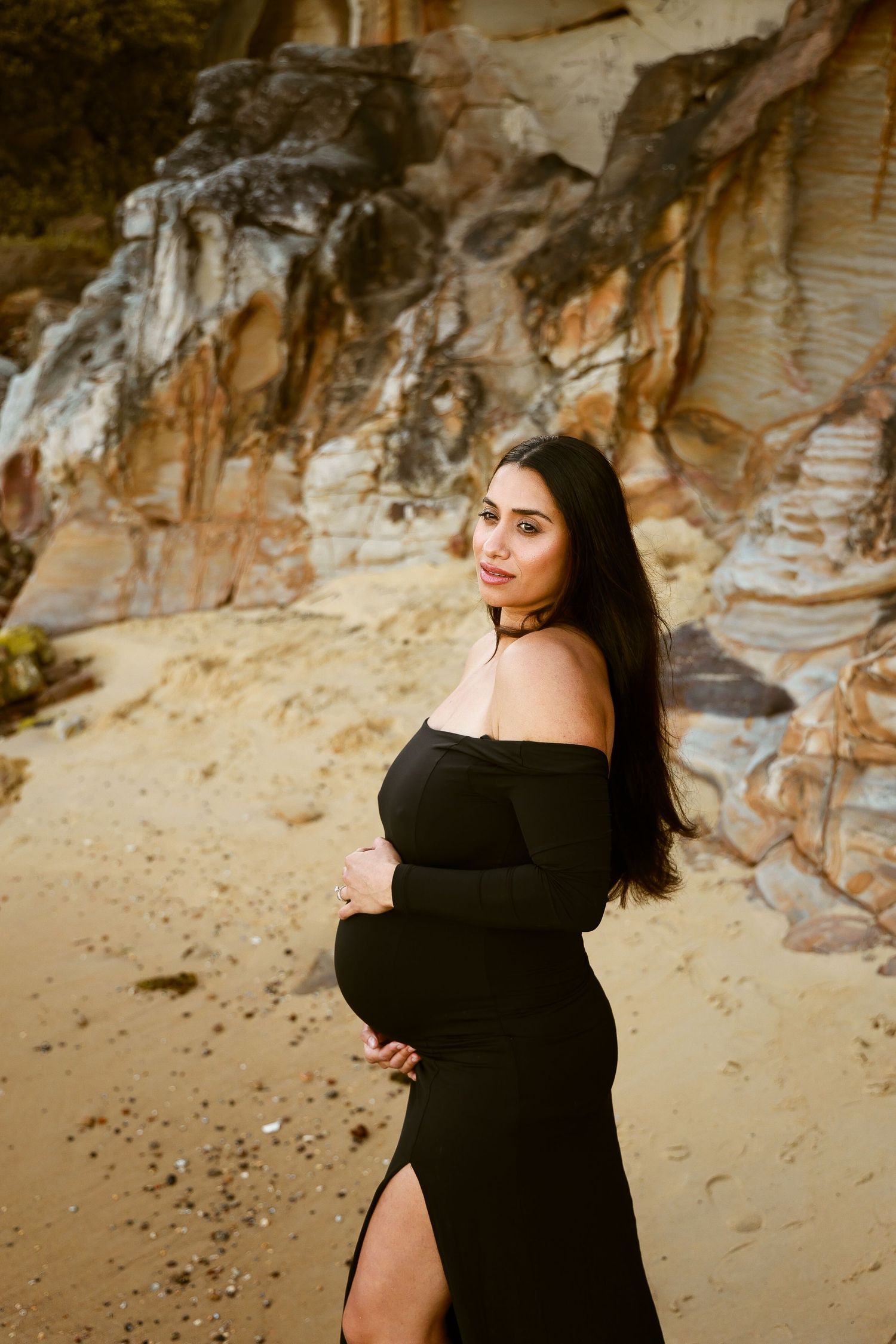 Maternity portrait of woman in black dress against dramatic coastal cliff backdrop.