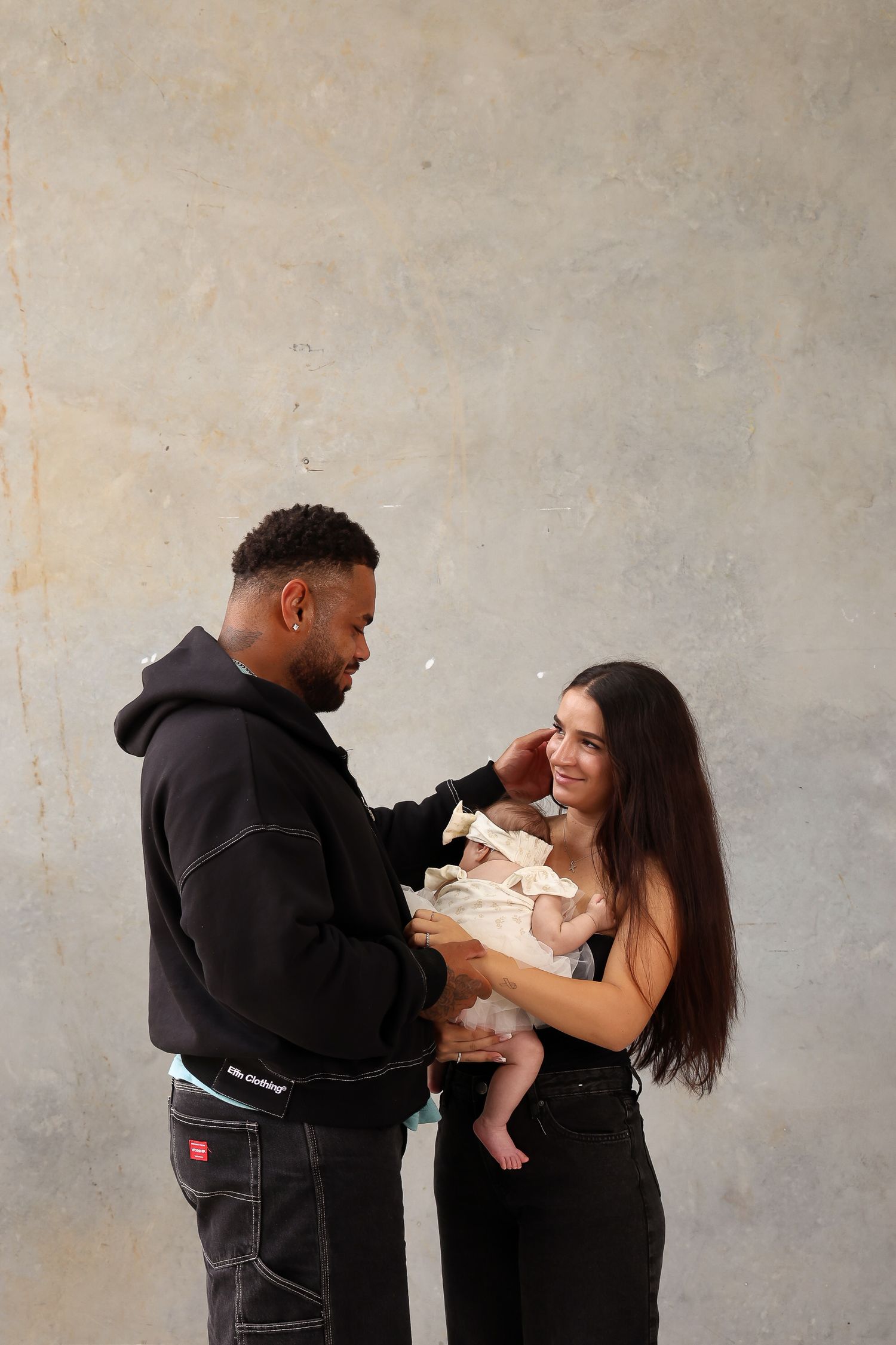 A couple shares a tender moment with their baby against a neutral gray background.