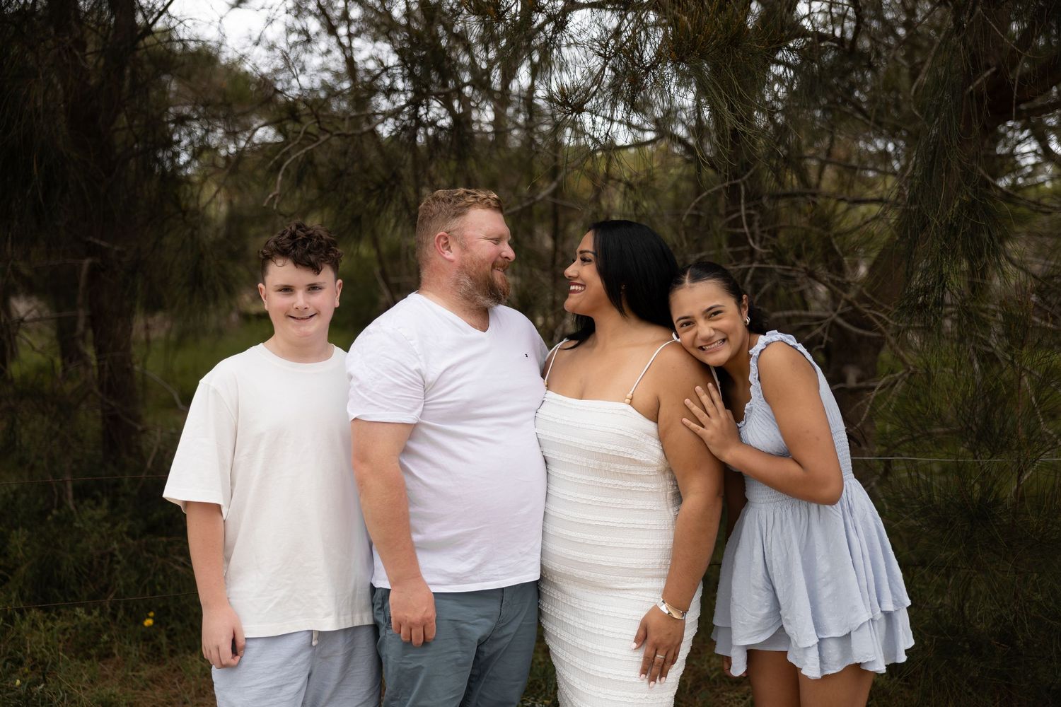 Family of four smiling outdoors among trees, wearing casual white and blue outfits during a professional photo session.