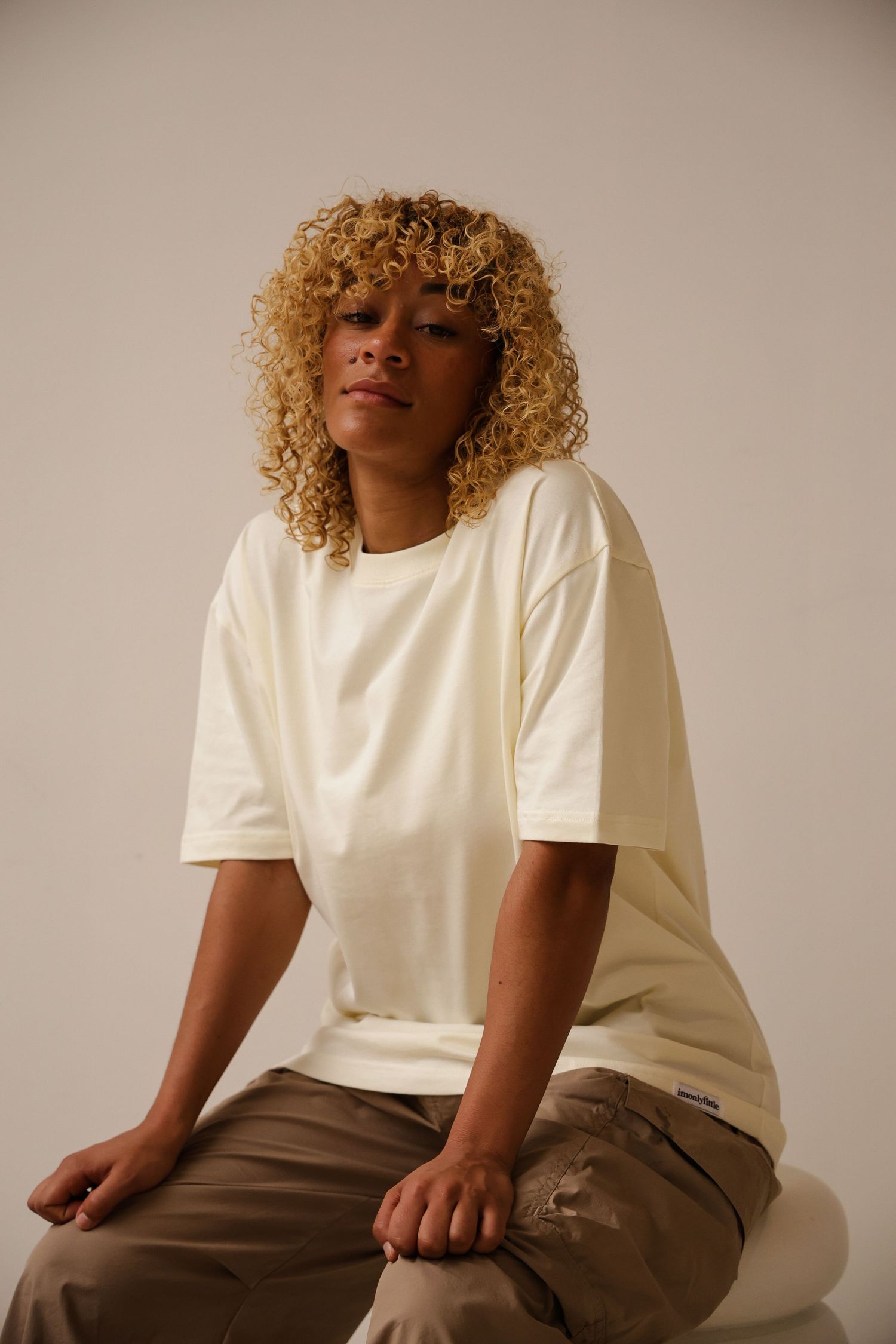 Person in white t-shirt and brown pants sitting against neutral backdrop with curly blonde hair.