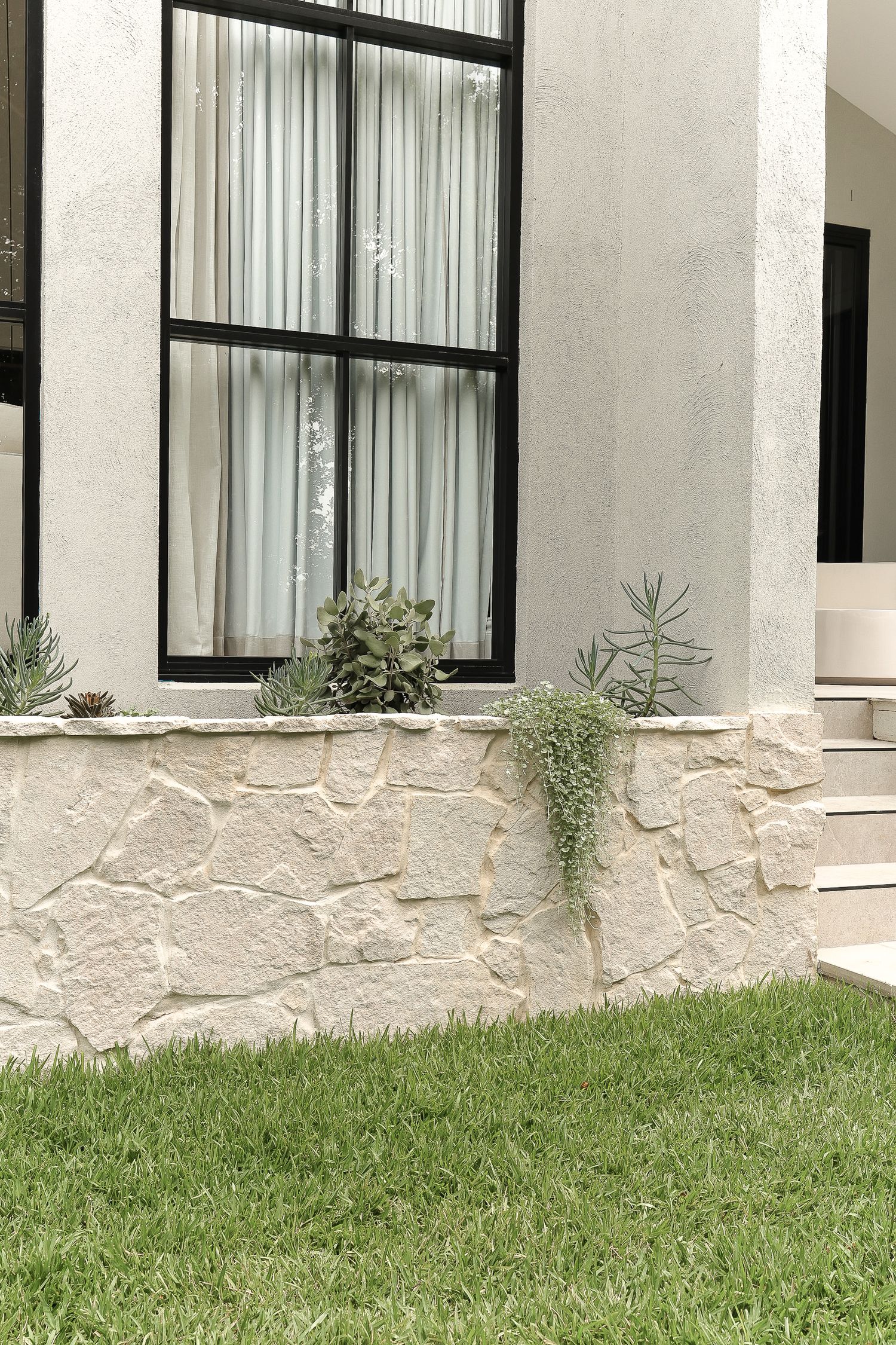 White exterior wall with large windows and decorative stone base featuring potted plants.