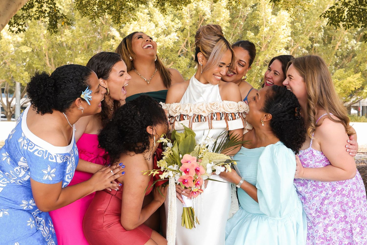 A joyful group in colorful dresses laughs together while gathered around a bouquet at an outdoor celebration.