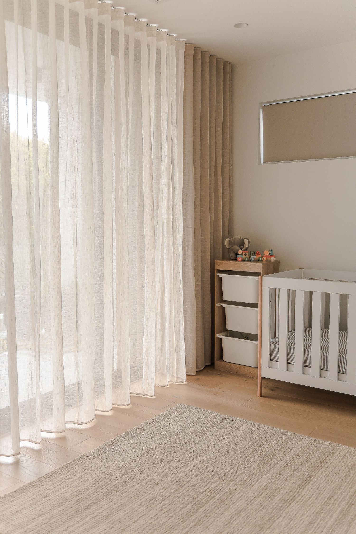 Nursery room with white curtains, wooden floor, and white crib beside a storage unit.