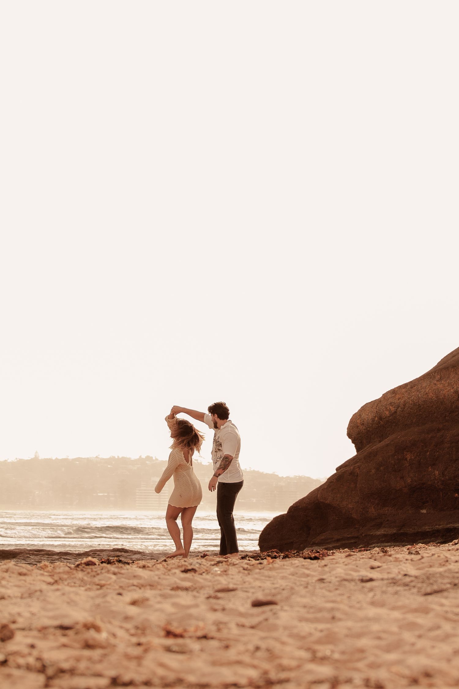 Romantic couple silhouetted against sunset on beach near large rock formation during golden hour.