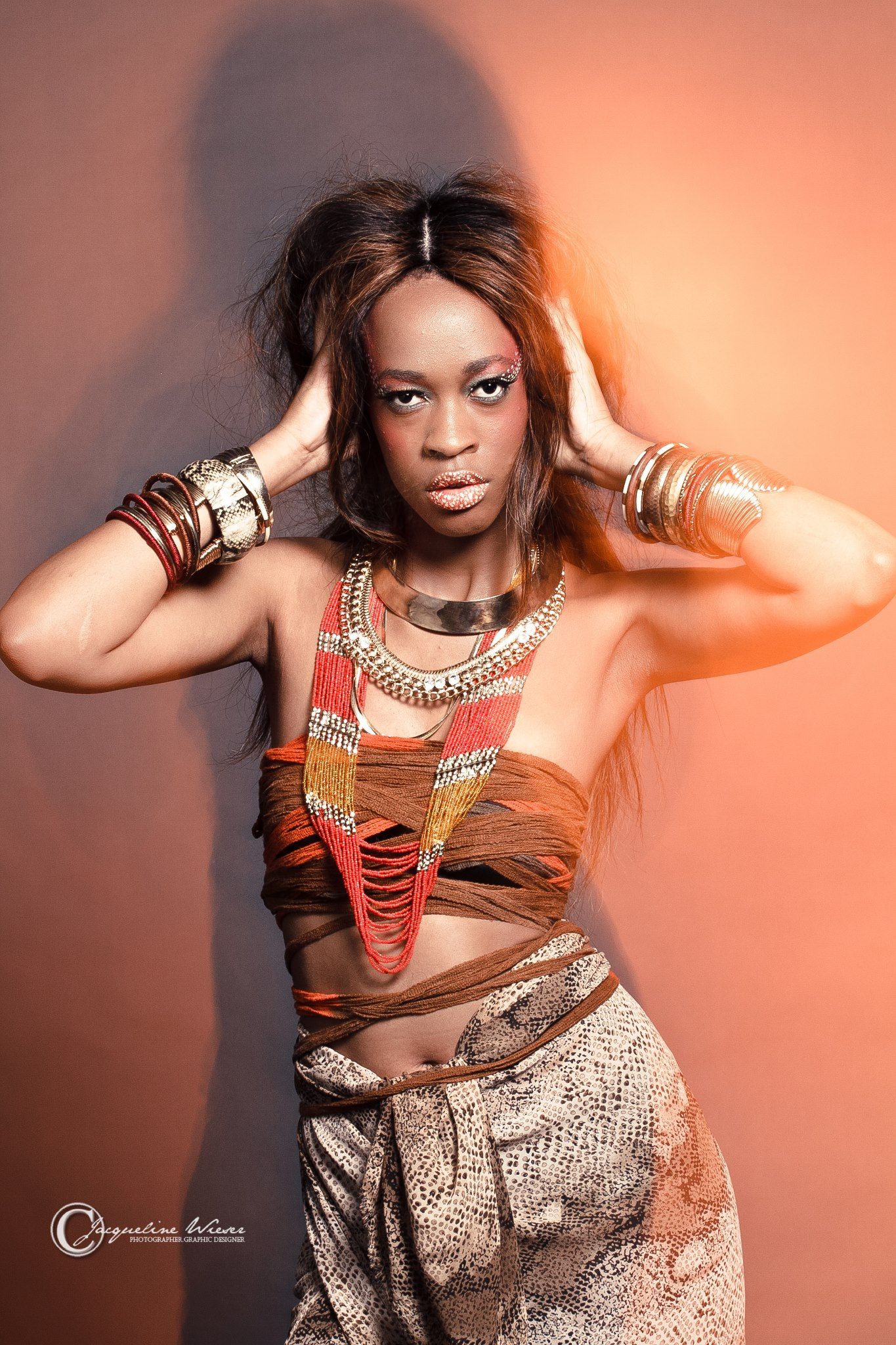 Model wearing tribal-inspired fashion with statement jewelry and crop top against orange background.