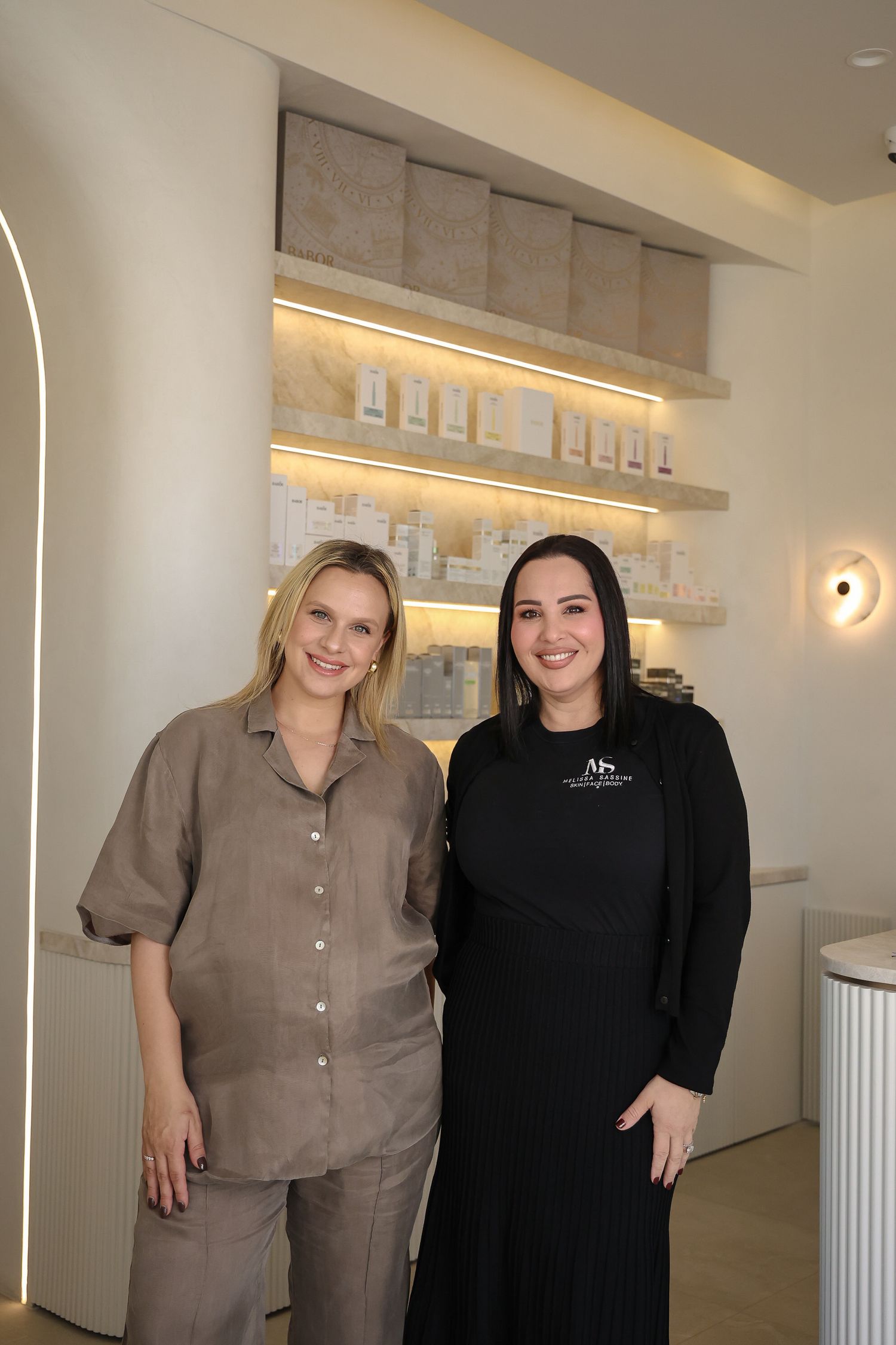 Two people stand in a modern spa or salon interior with illuminated shelving and minimalist decor.