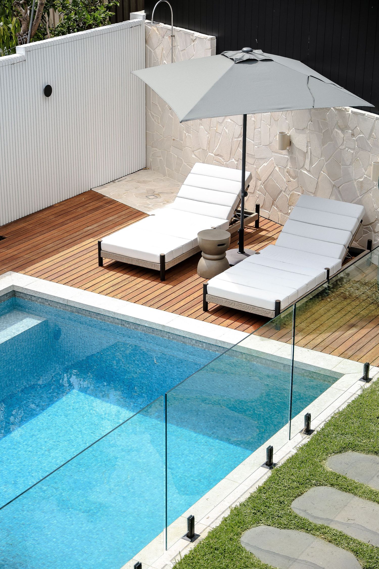 Poolside deck with two white loungers and umbrella beside a glass-fenced swimming pool.