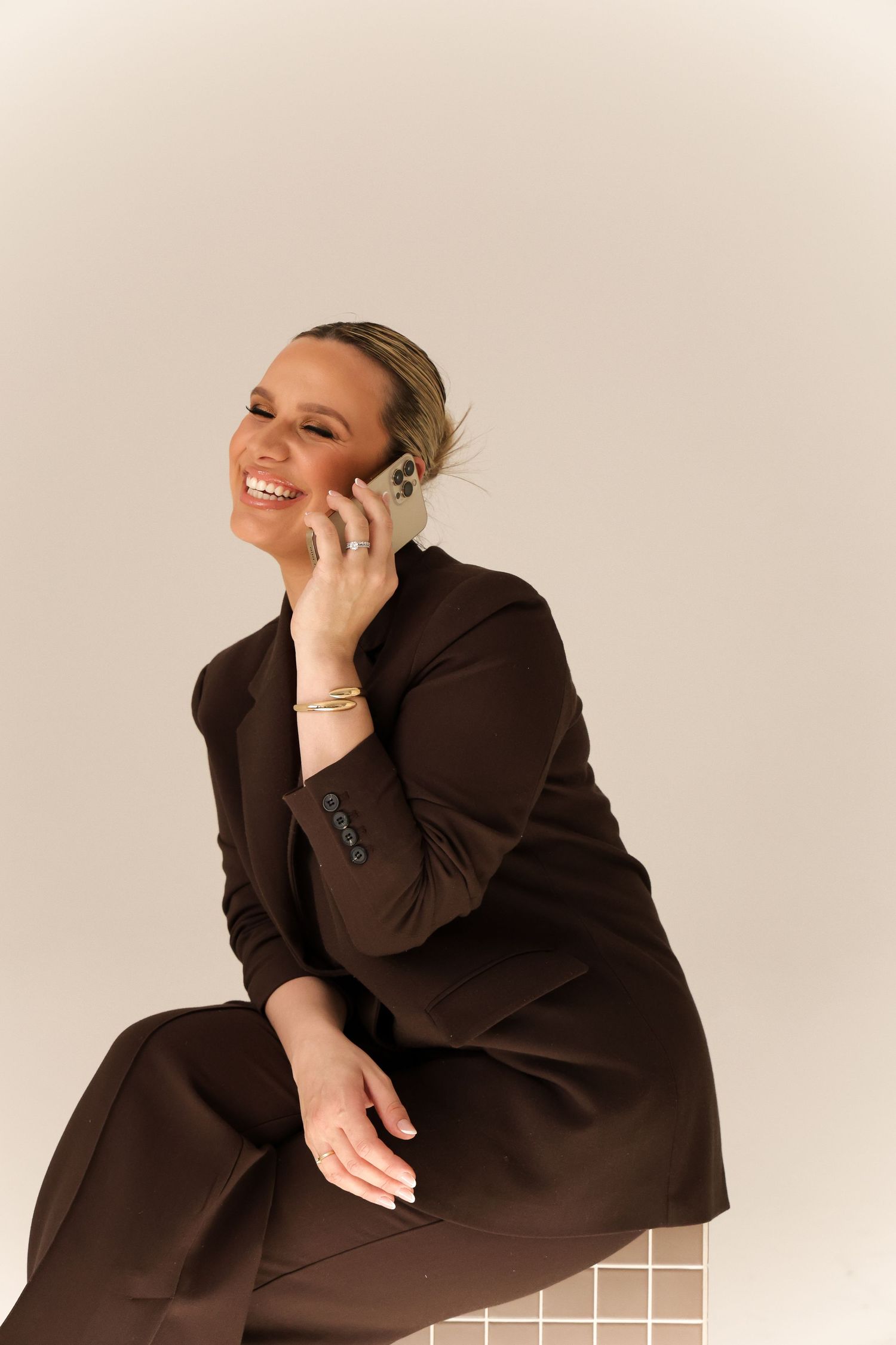 Person in dark brown business attire smiling while talking on phone in studio setting.