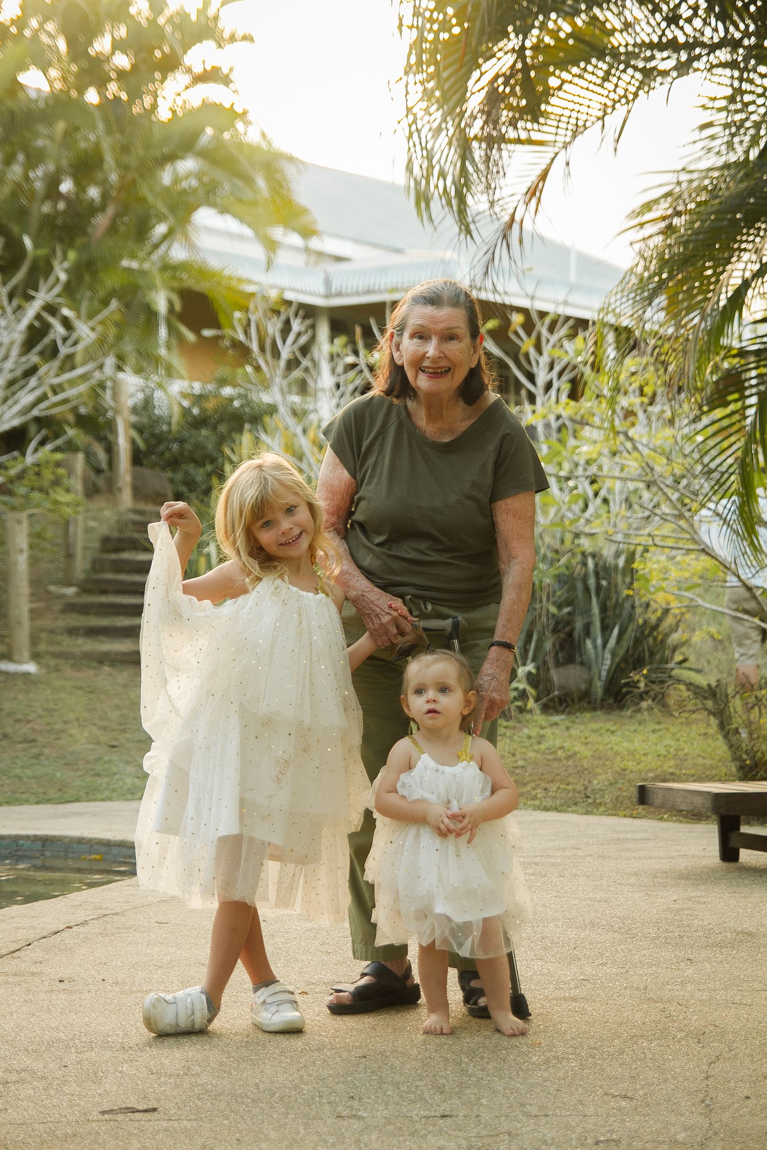 Three people in white dresses pose together on garden path surrounded by palm trees.