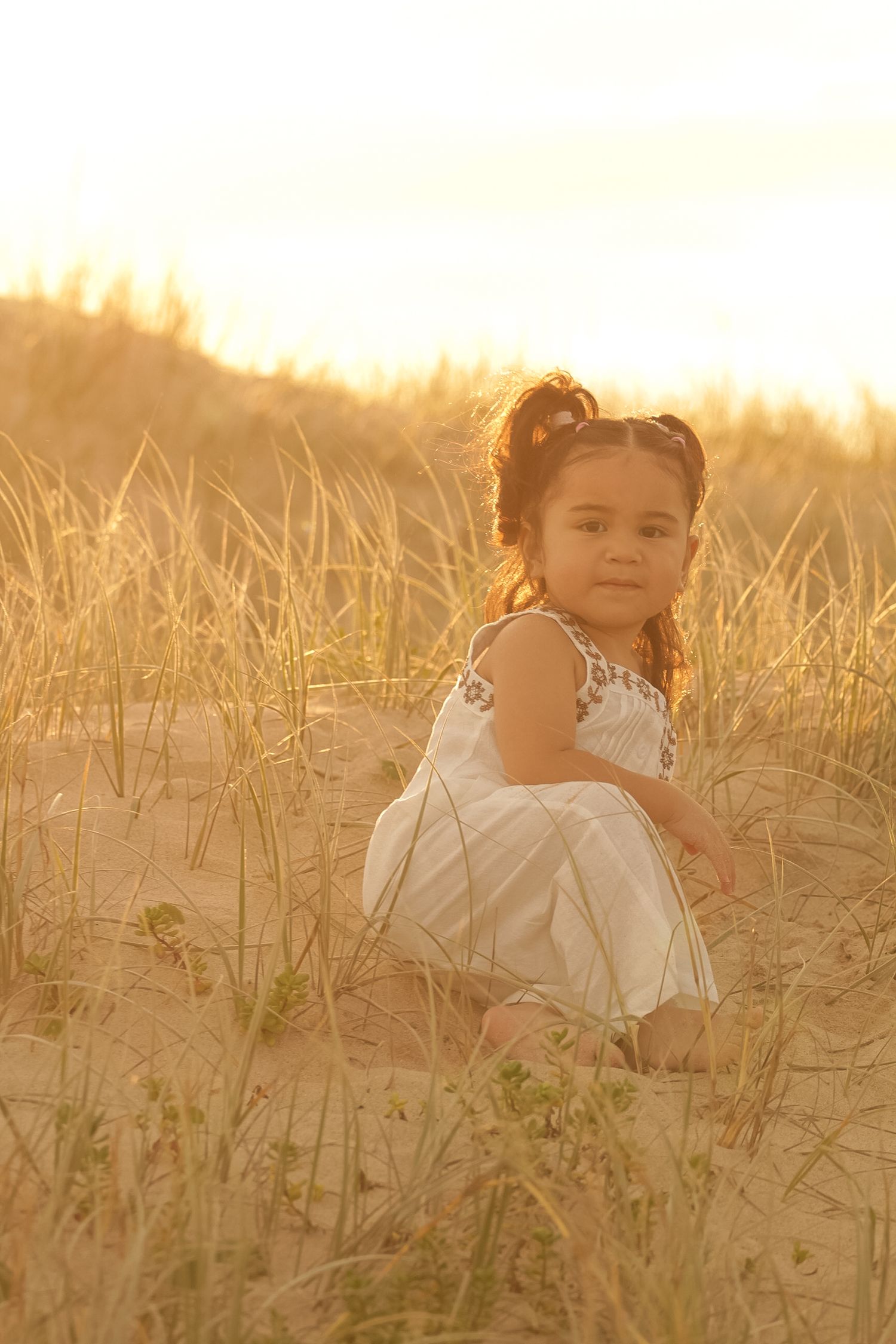 A white dress-clad figure sits in tall grass during golden hour sunset.