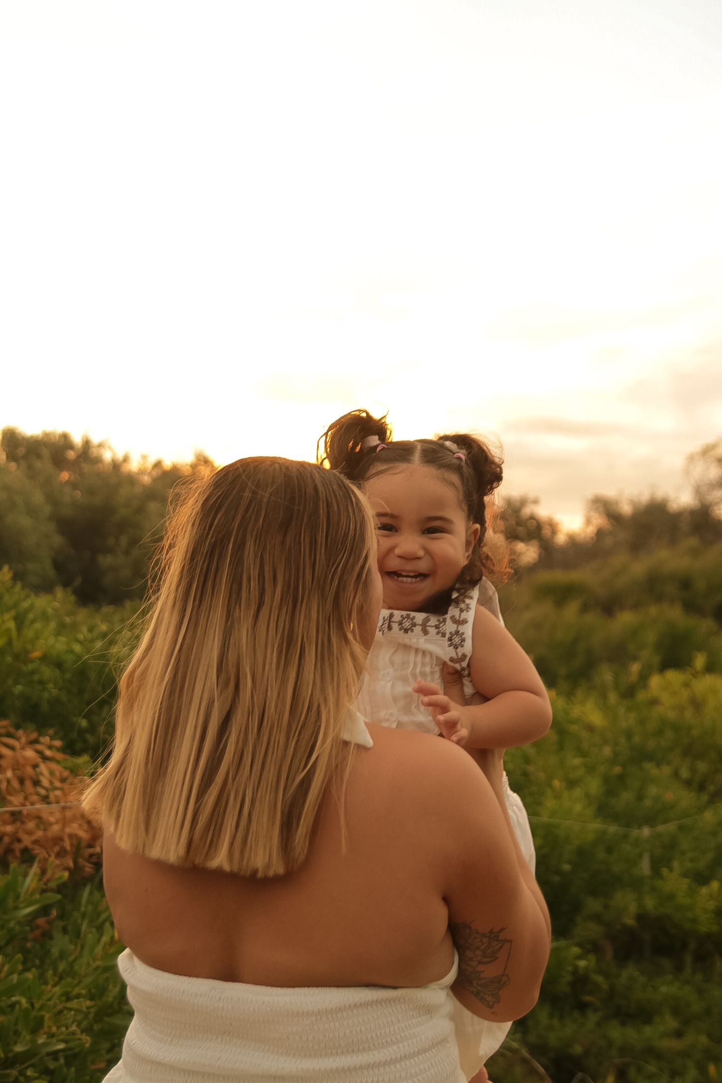 Parent and child share a tender moment at sunset among coastal greenery.