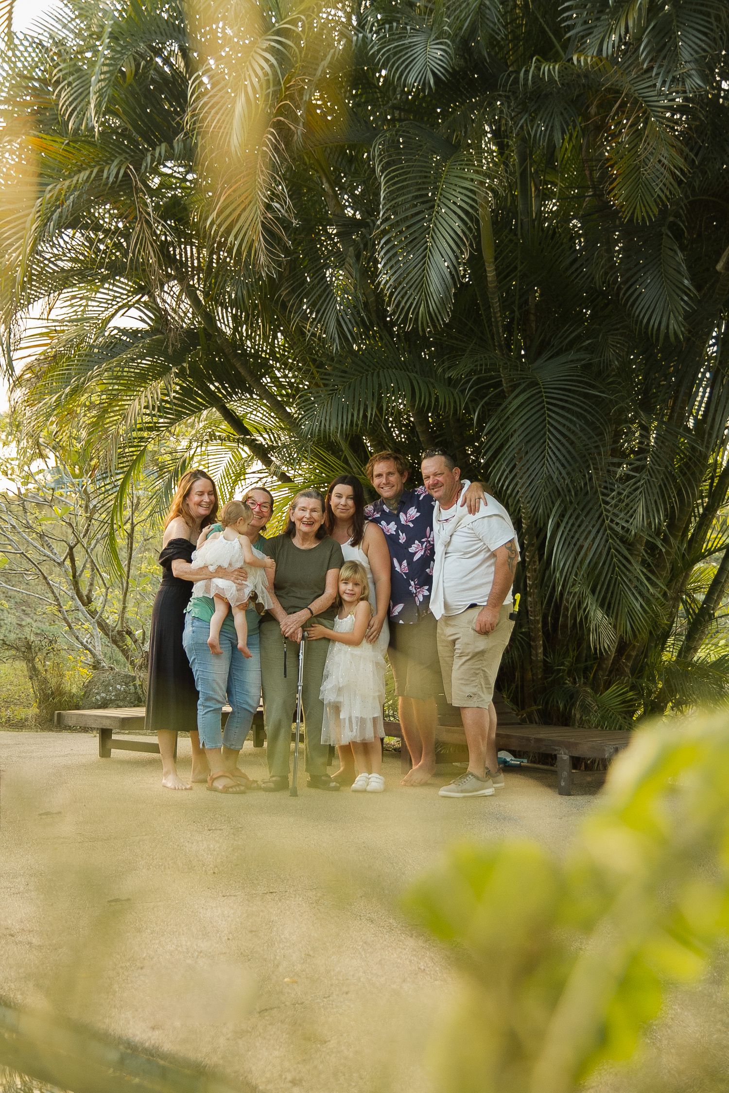 Multi-generational family poses together against lush palm trees in tropical setting.