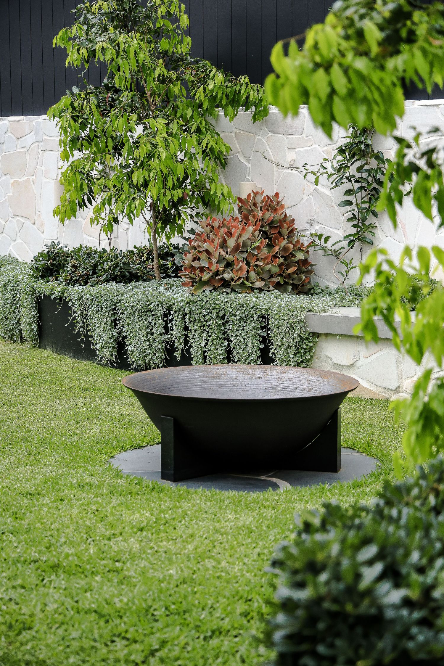 Large black fire pit bowl in a manicured garden with boxwood hedges and ornamental trees.