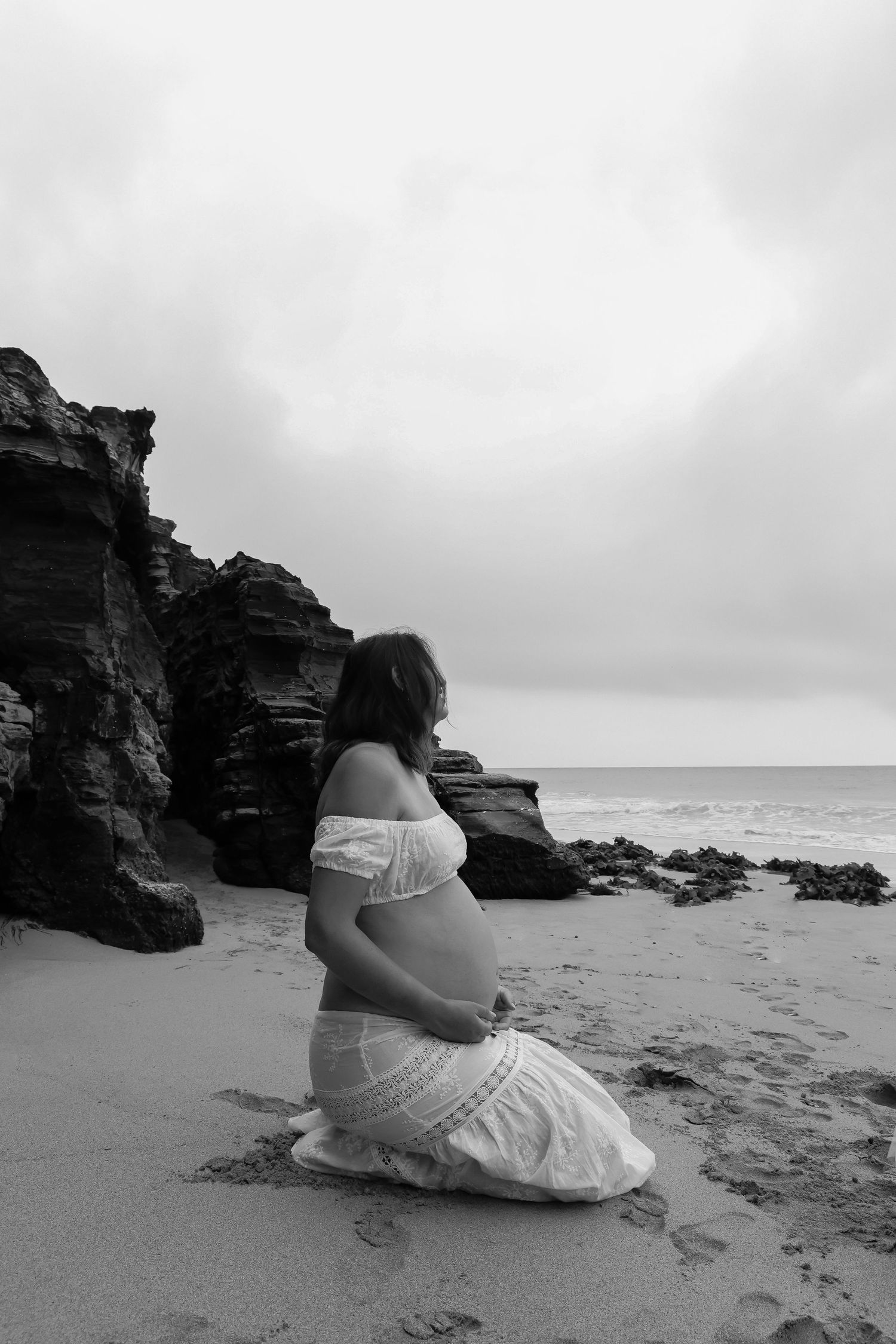 Person in white dress sitting on beach near rocky cliffs during overcast day.