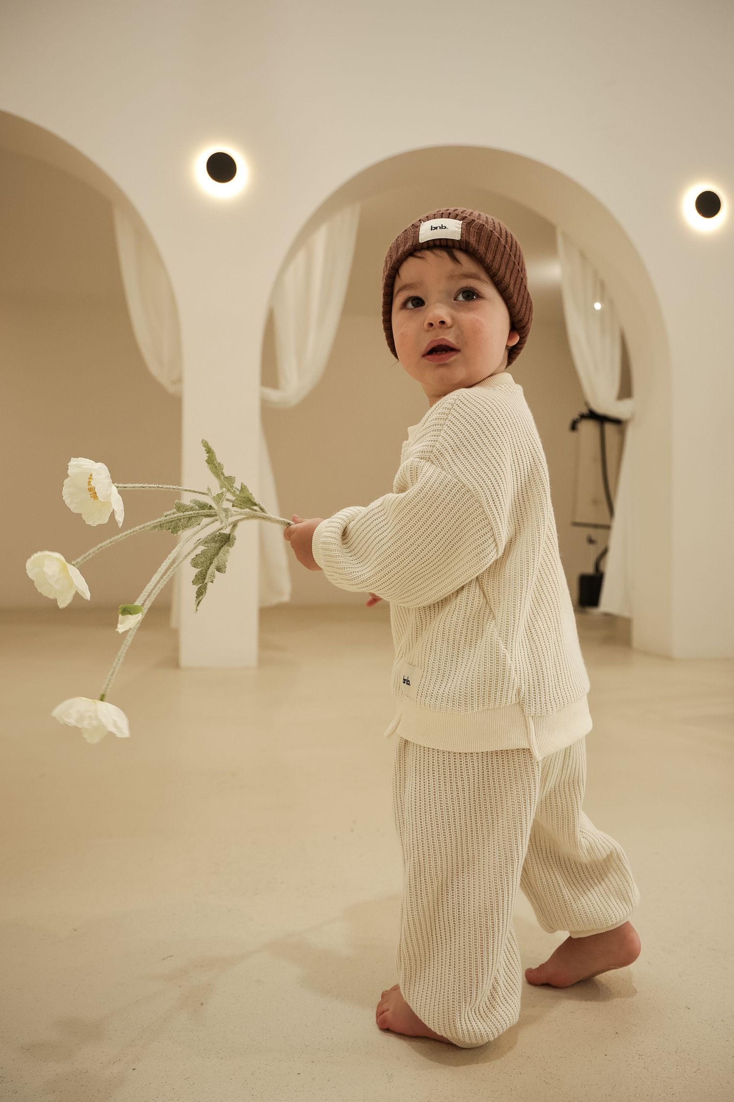 Baby in cream knit outfit playing with white flowers in a modern minimalist space.