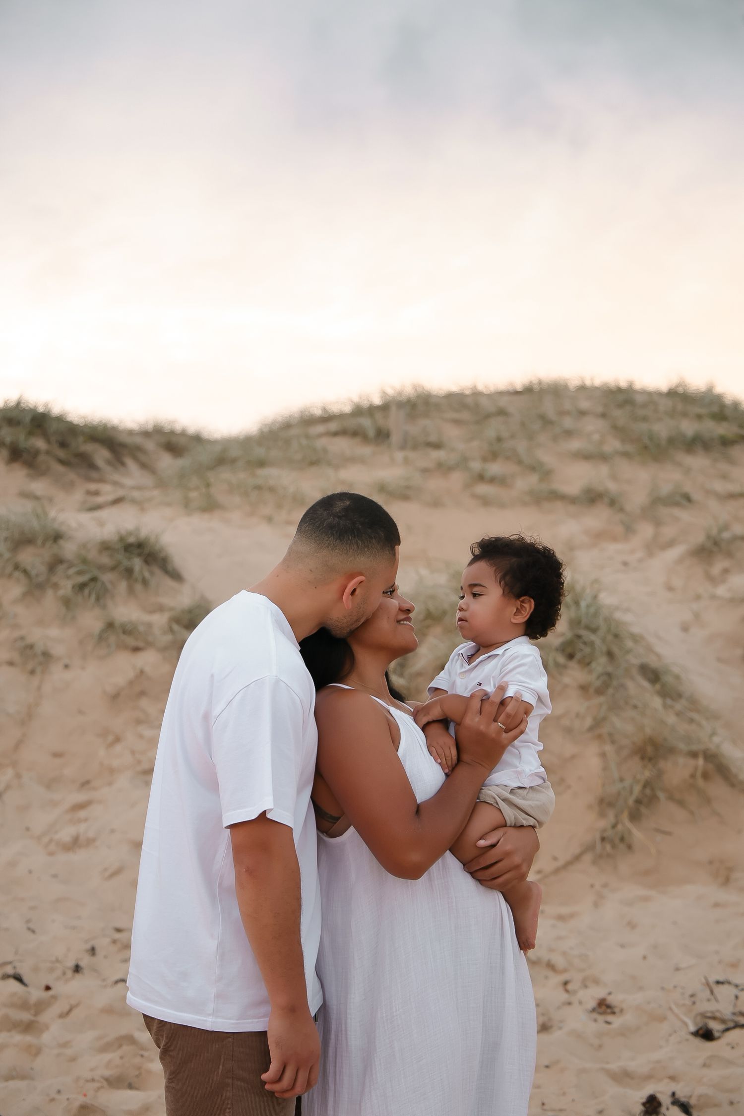 A tender family moment captured against sandy dunes at sunset.