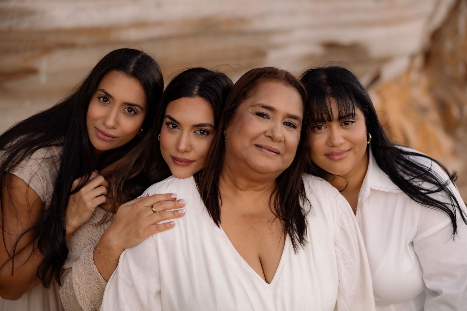 Four women in white tops pose closely together smiling outdoors against a natural stone background.
