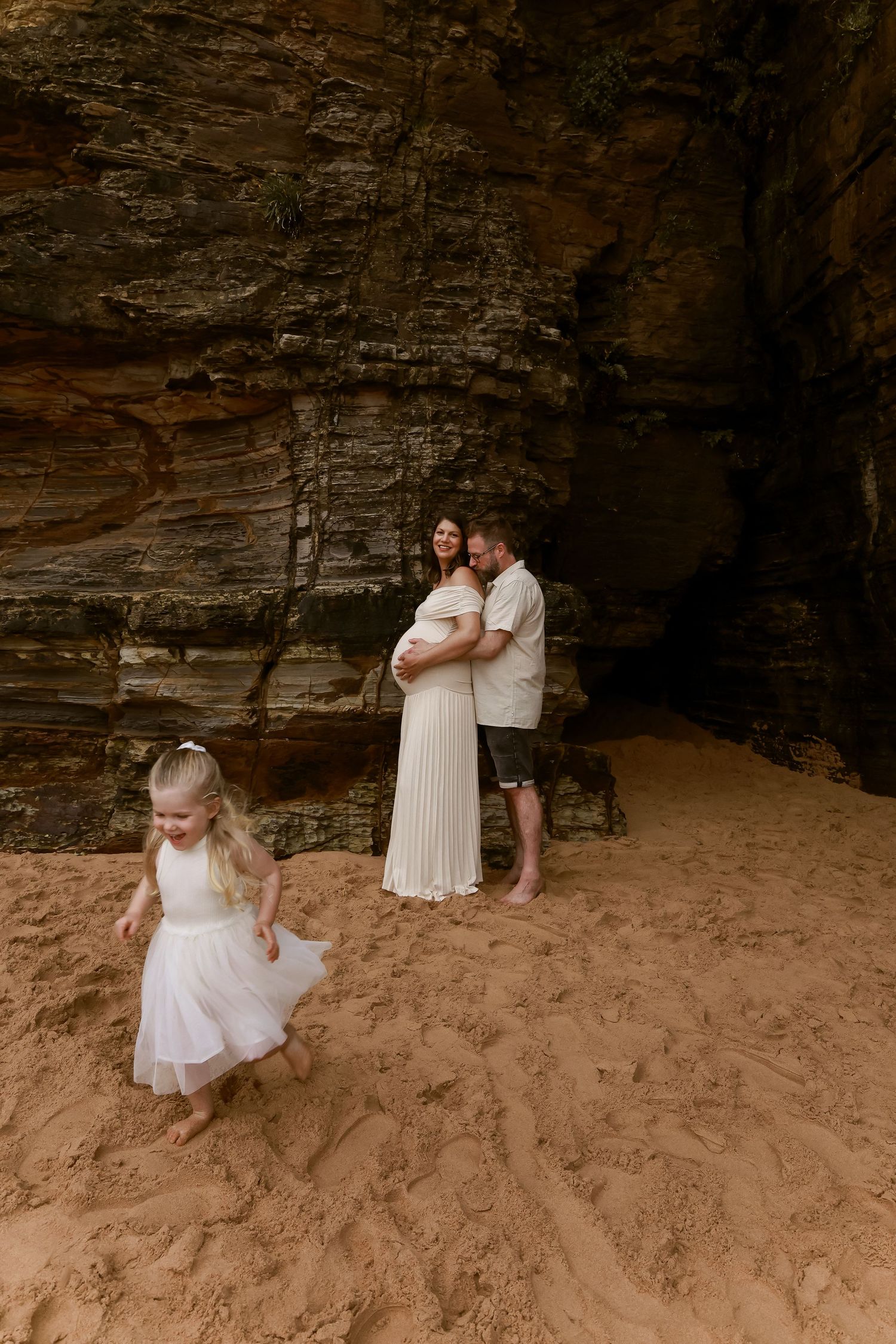 A family maternity photo at a rocky beach cave with a small child playing in the sand.