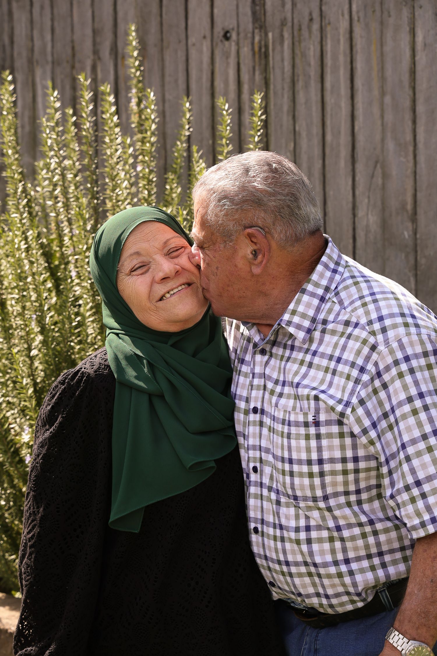 Candid moment of affection between elderly couple by rustic wooden wall with flowering plants.