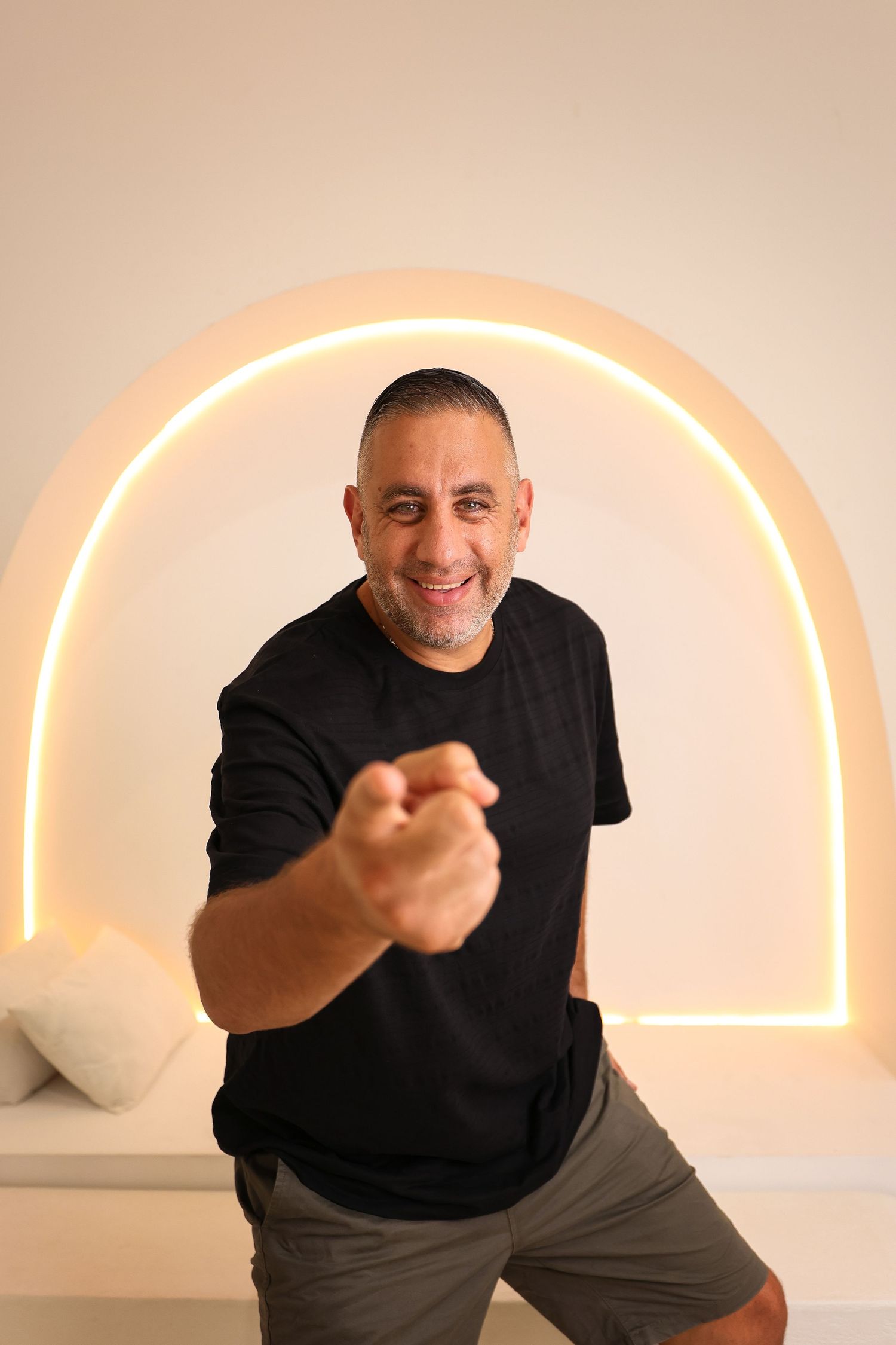 Person in black t-shirt pointing at camera with illuminated arch in background.