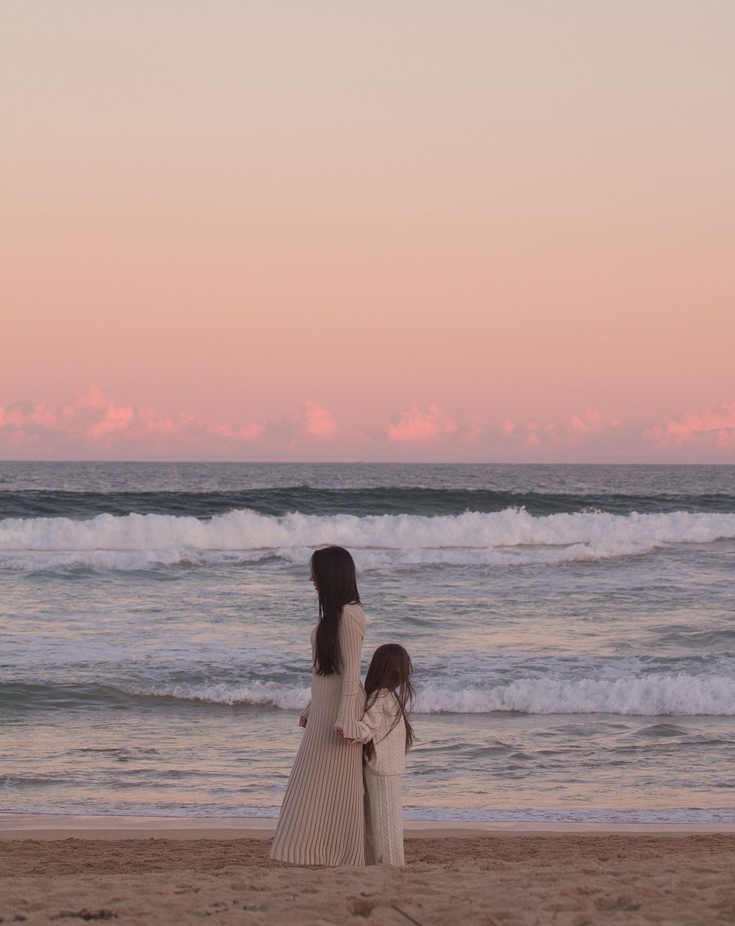 Two figures sit together on the beach watching waves roll in during a pink sunset.