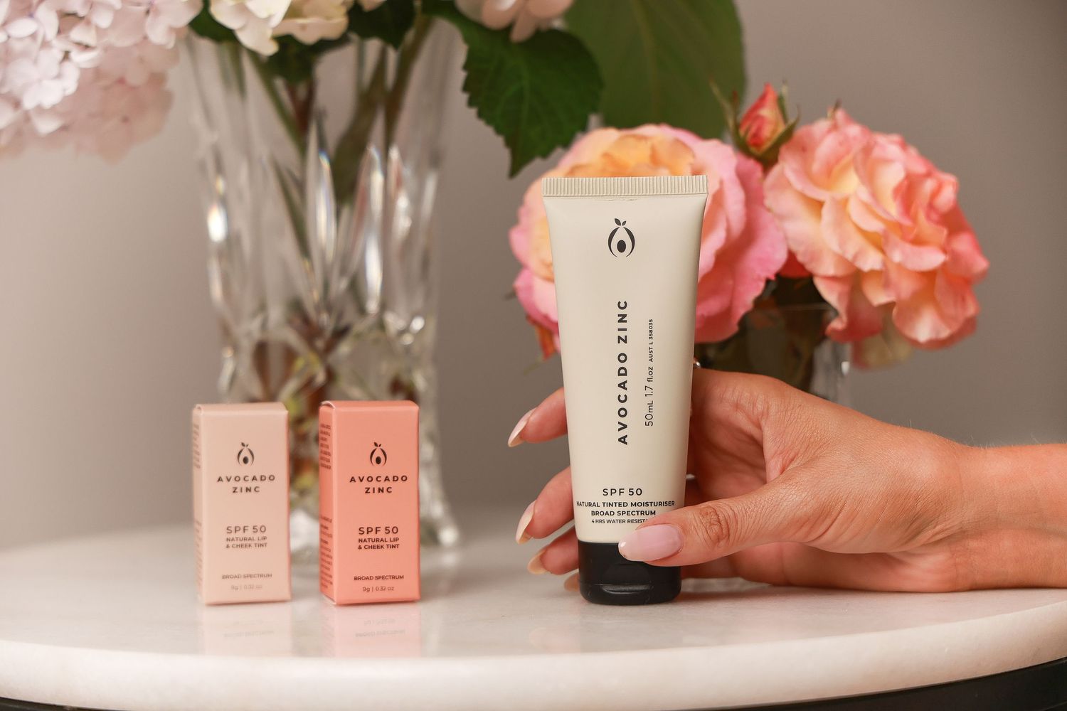 Beauty products displayed with pink flowers and crystal glassware.