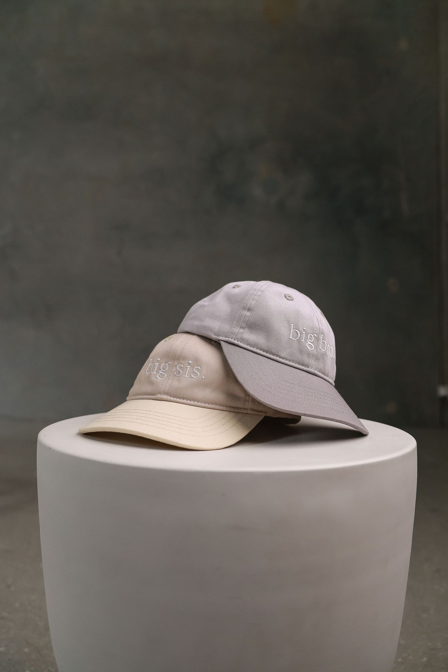 Three light colored baseball caps stacked on a white cylindrical pedestal.