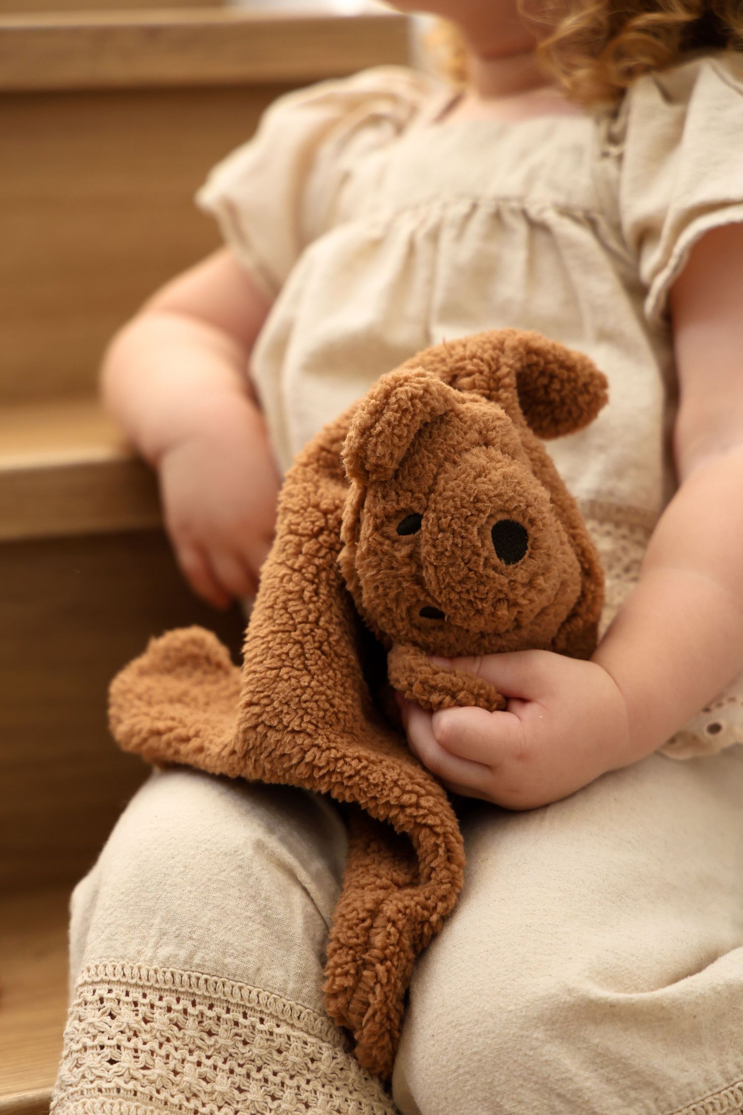Small hands holding a plush brown stuffed bunny toy against a cream-colored dress.