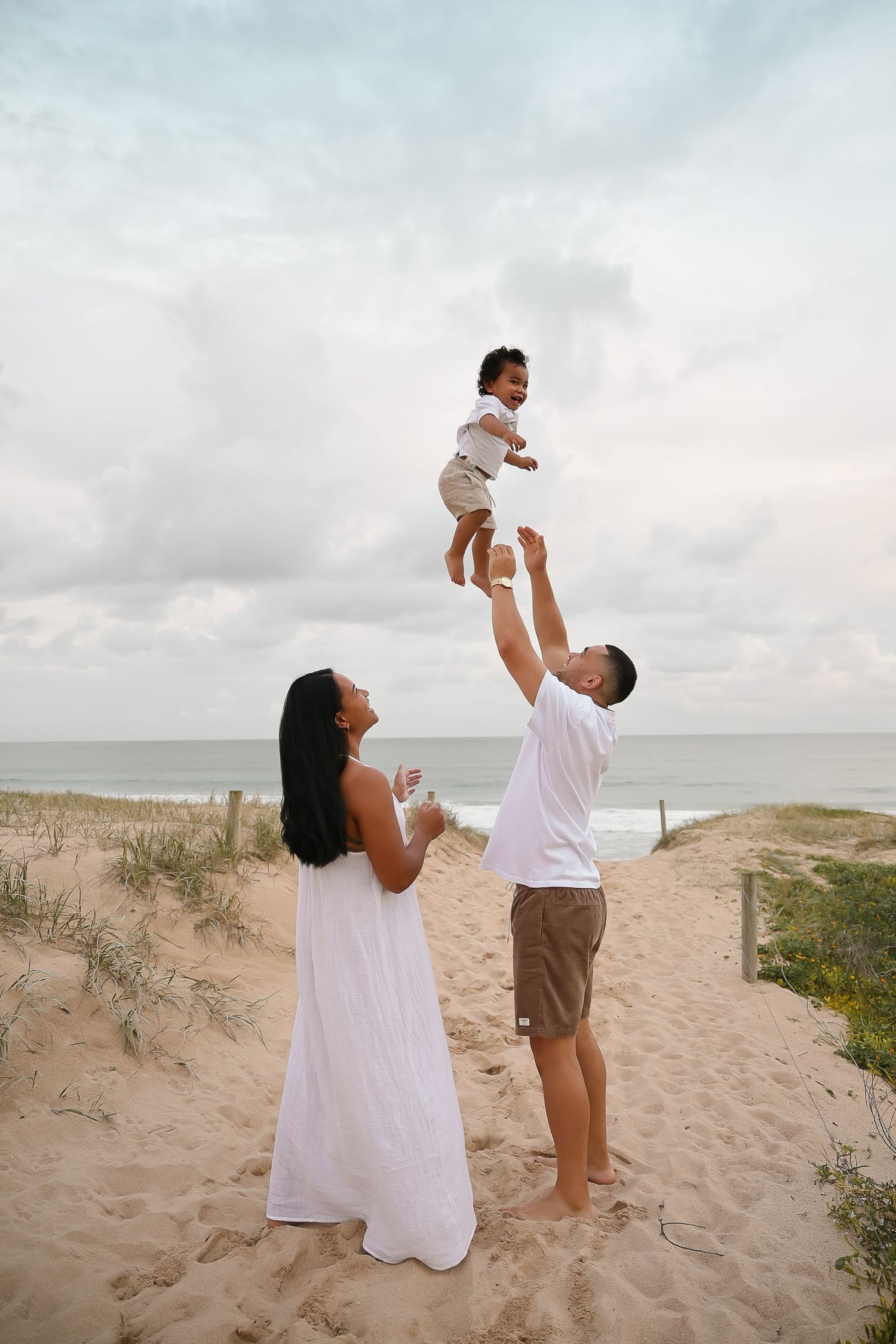 Family plays together on sandy dunes with ocean backdrop during golden hour.