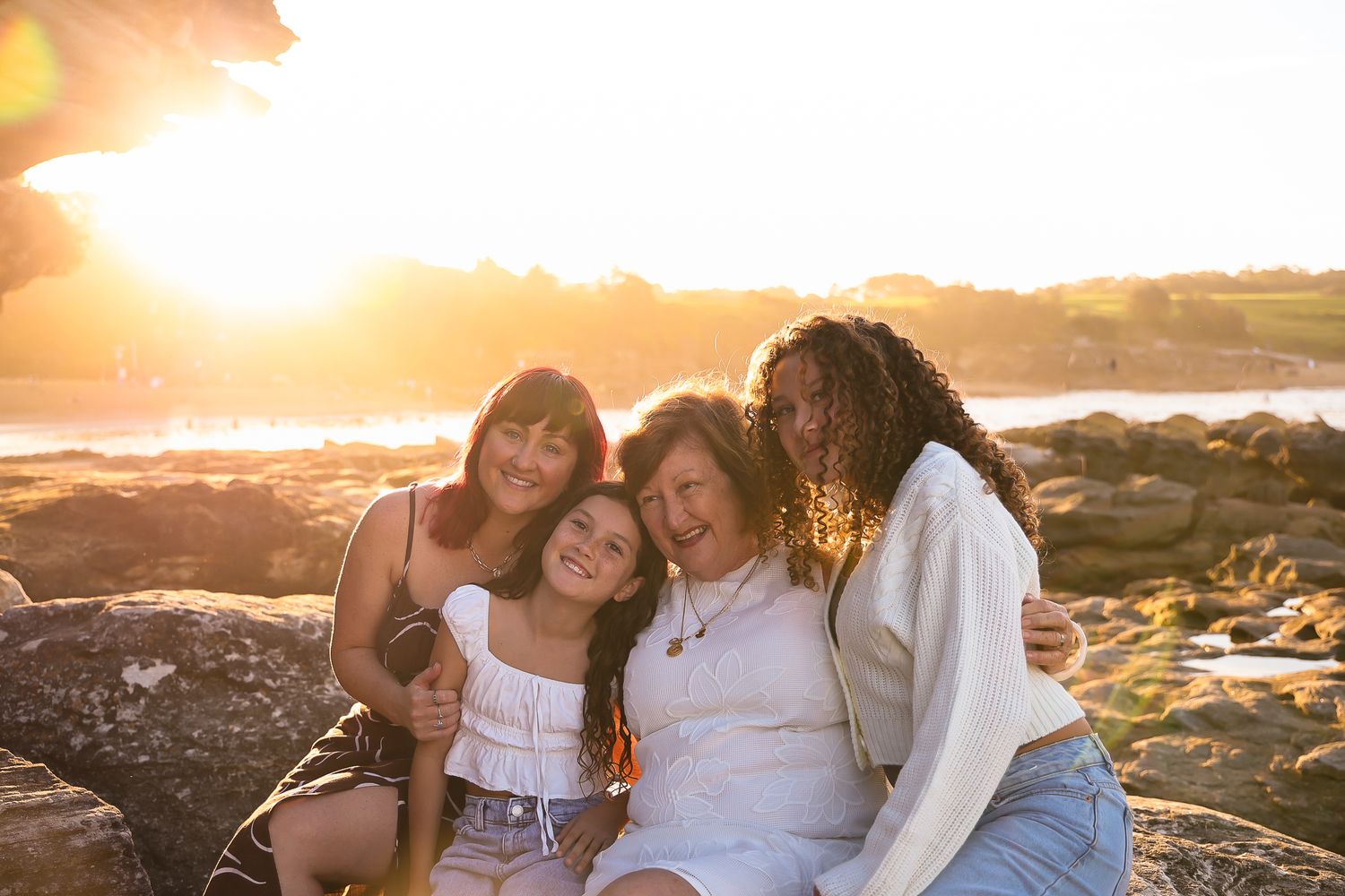 A family sits together on rocks at the beach during golden hour sunset with waves crashing in the background.