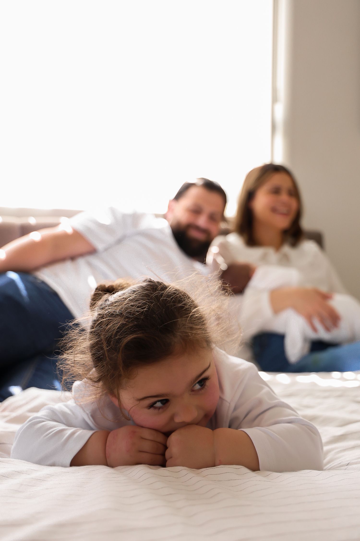 A young girl lies on a white bed resting her chin on her hands while her smiling parents relax together in the background.