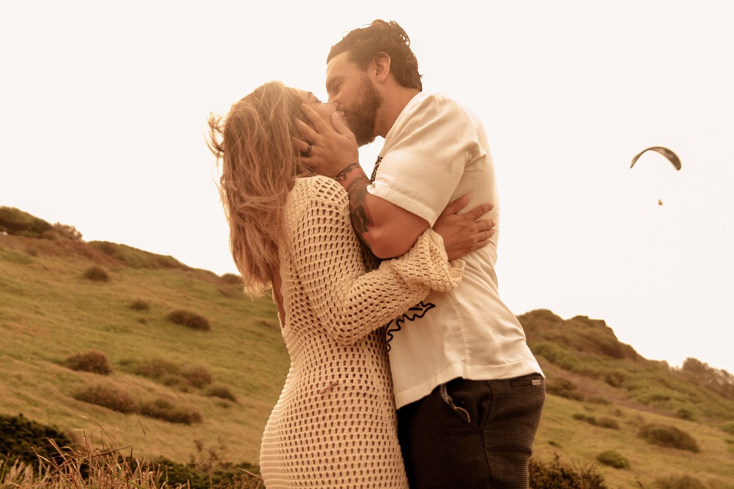 Romantic couple embracing on hillside with paraglider in background.