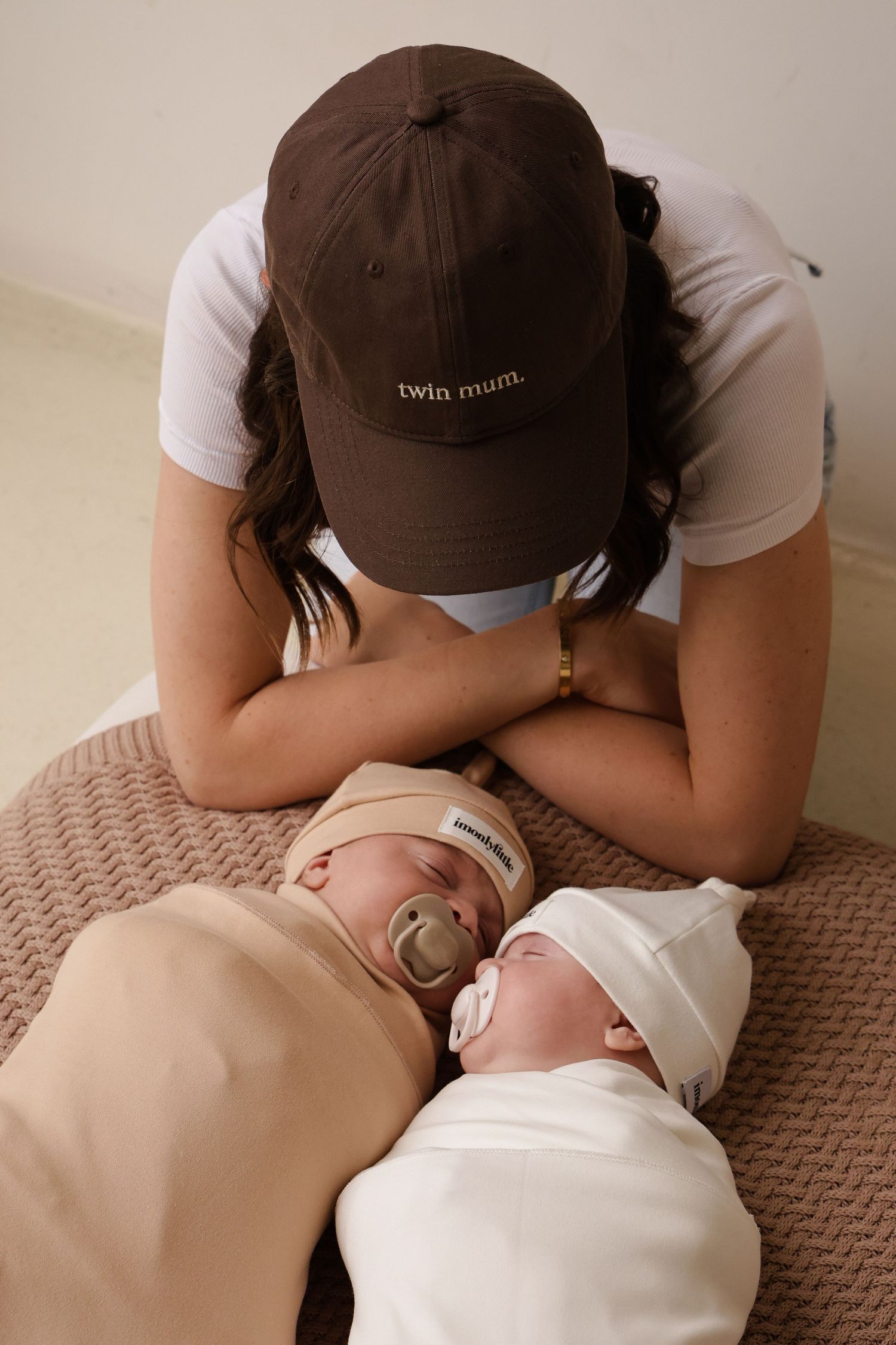 A parent wearing a brown 'twin mom' hat leans over two sleeping infants on a textured blanket.