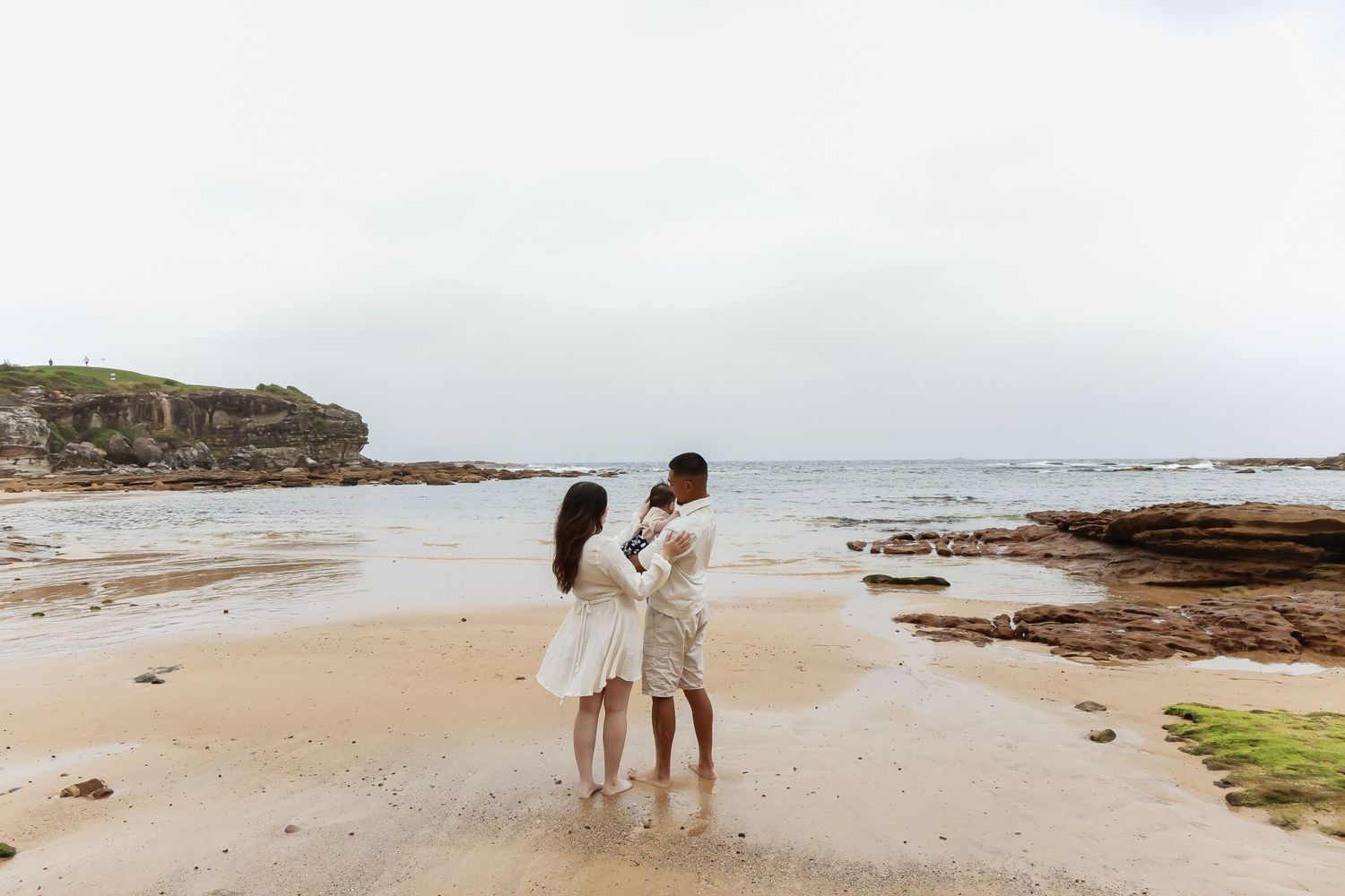 Couple embracing on rocky beach coastline with cloudy sky and ocean waves in background.