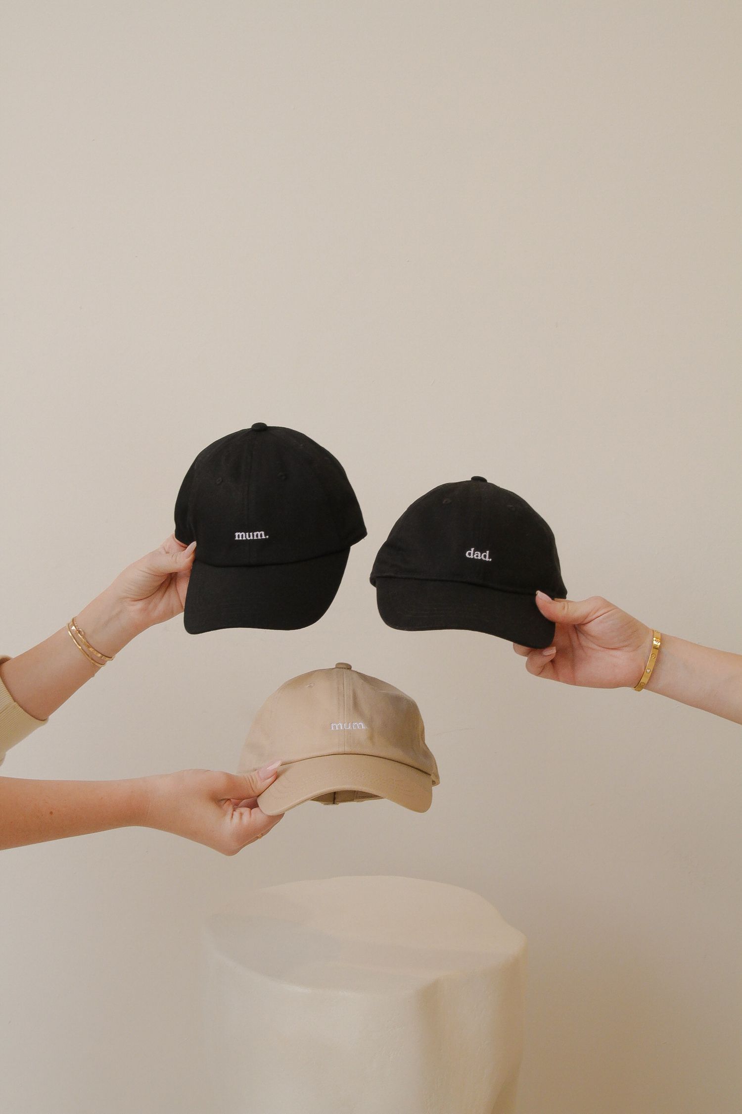 Hands holding three baseball caps in black and beige colors against neutral background.