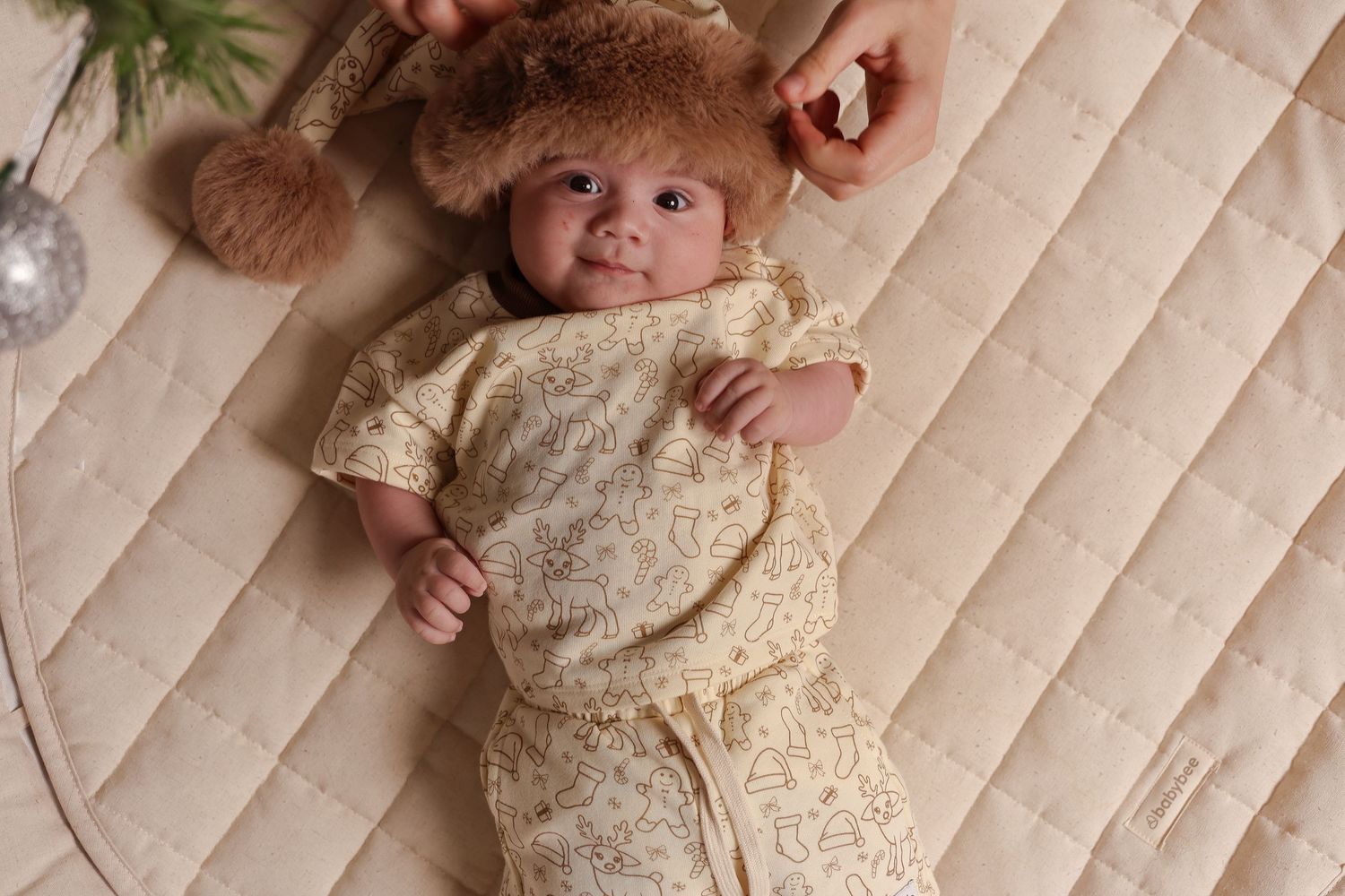 Baby wearing cream colored outfit with furry winter hat lying on quilted surface.