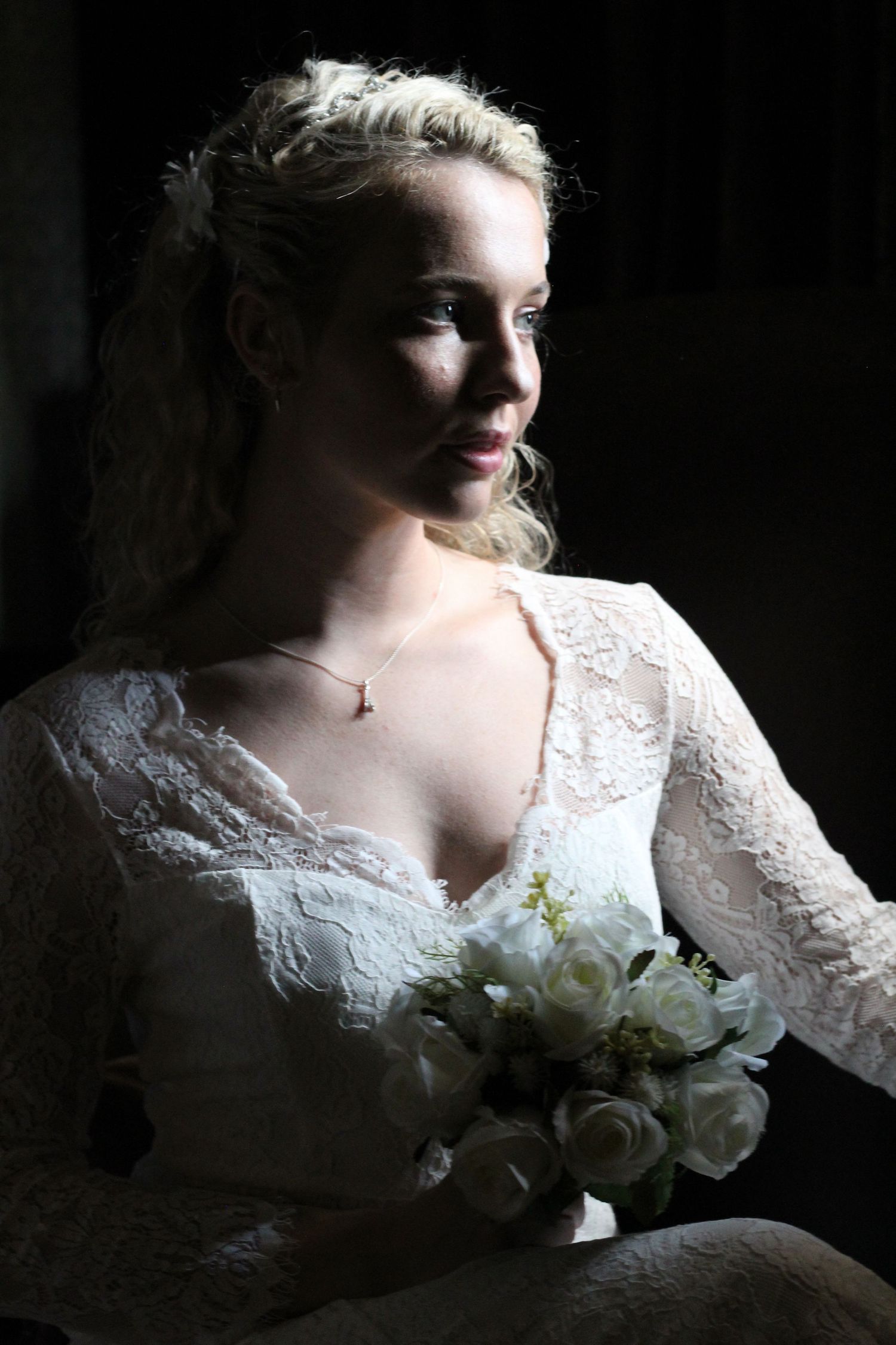 Close-up portrait of a bride in white lace dress holding a white bouquet in dramatic lighting.