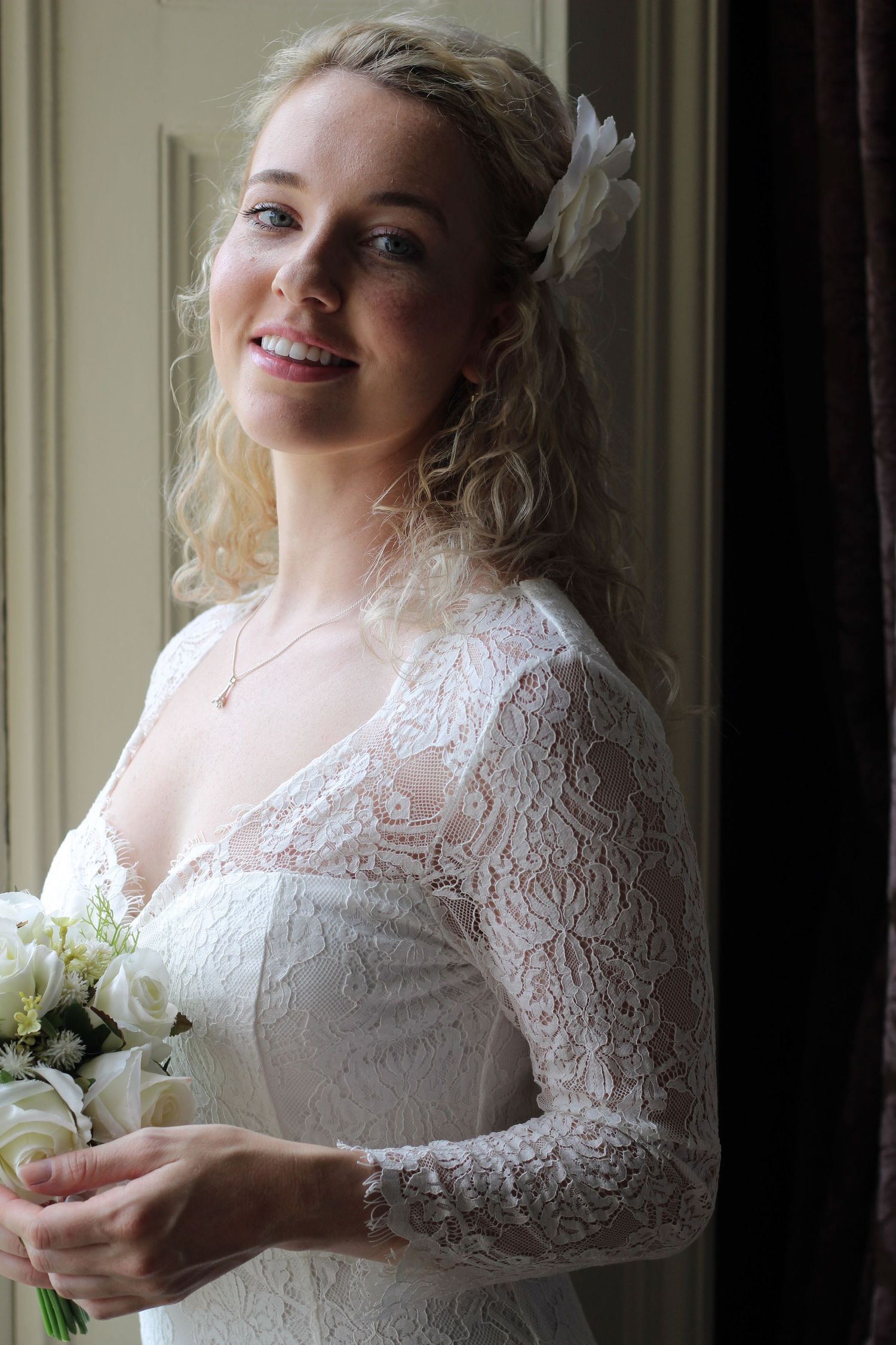 A bride in a lace long-sleeve wedding gown poses by a window with a white bouquet.
