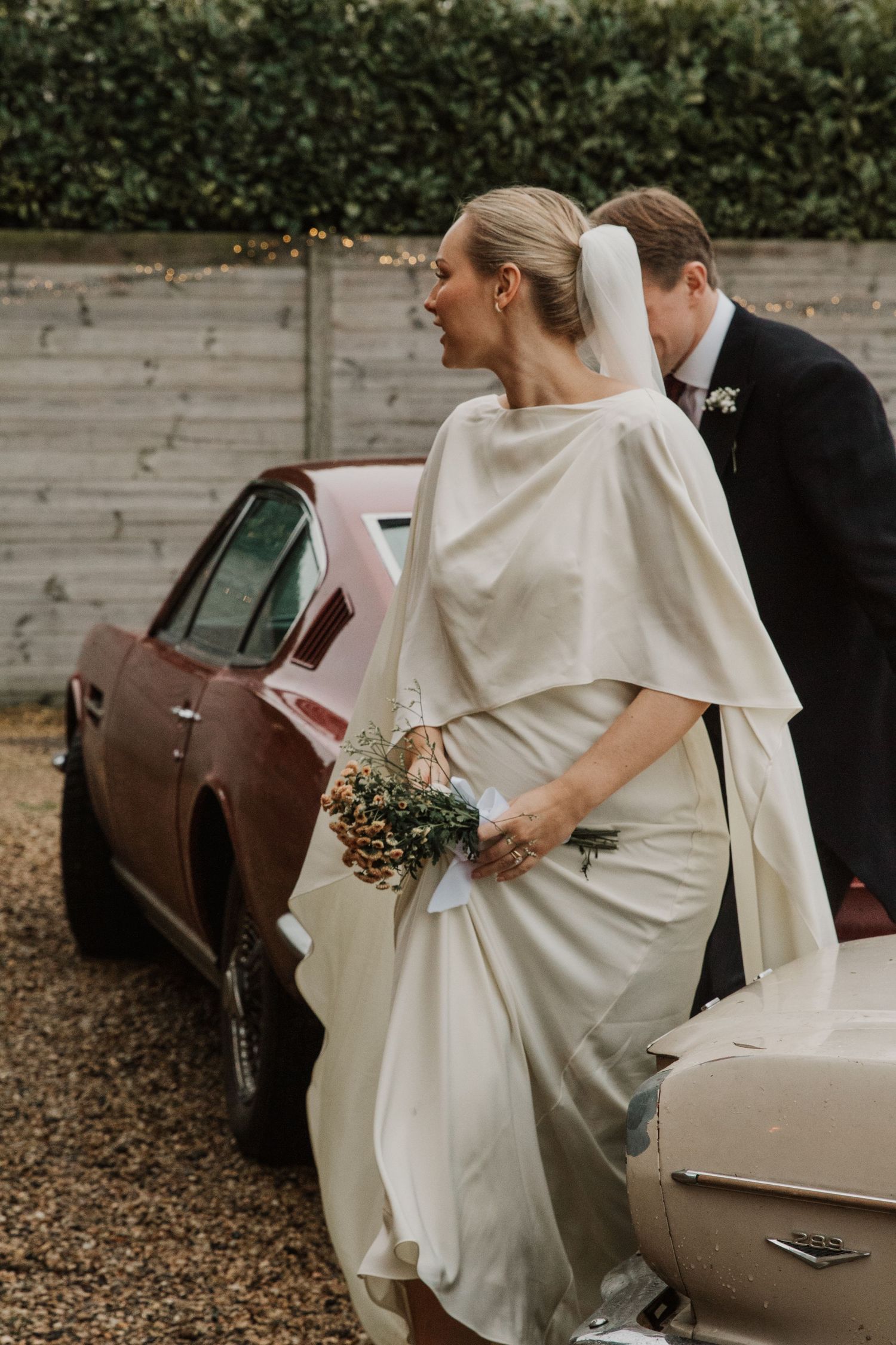 Elegant wedding couple next to a vintage pink car with the bride holding a bouquet.