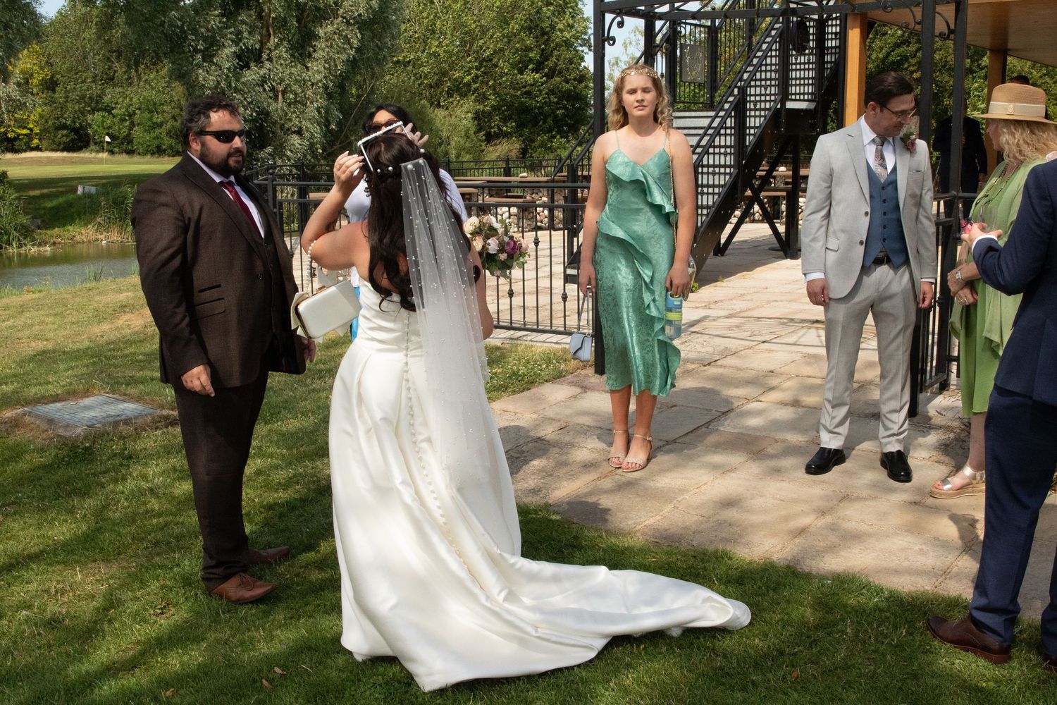 Wedding party gathered outdoors on a sunny day with guests in formal attire and a flowing white wedding gown.