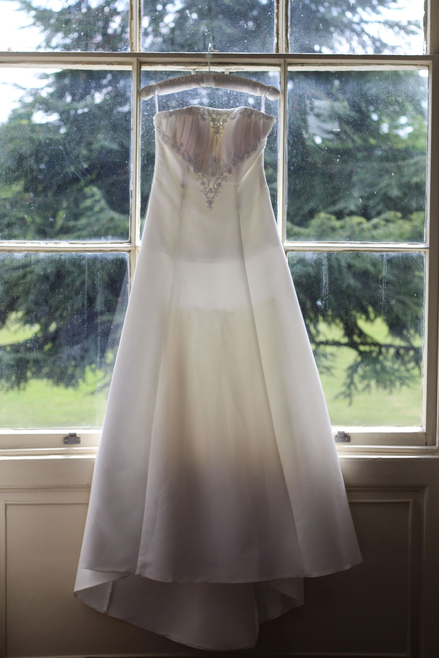 A white wedding dress with intricate beading hangs elegantly in front of sunlit windows.