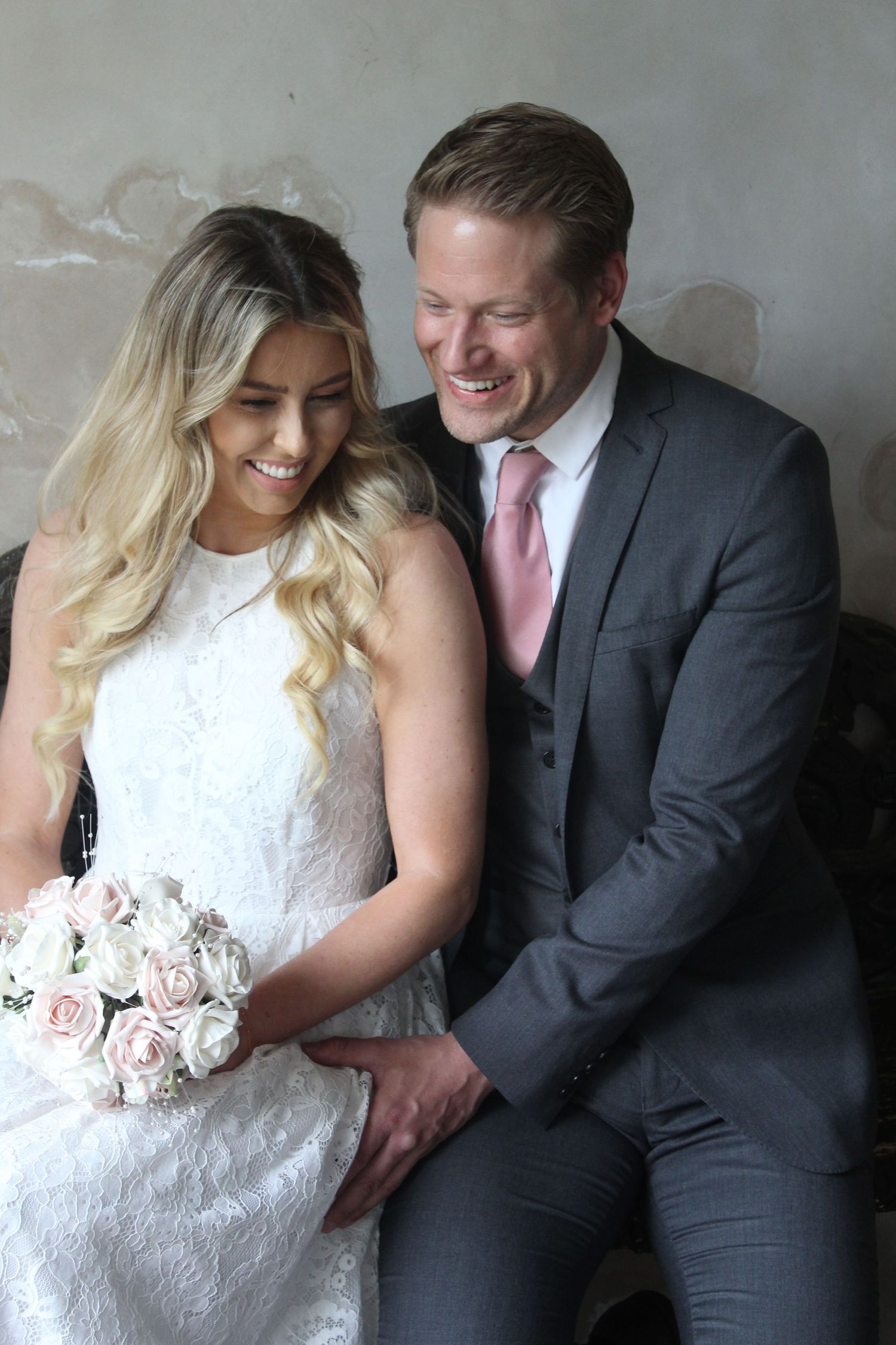 A romantic wedding portrait of a couple in formal attire with a pink rose bouquet against a grey background.