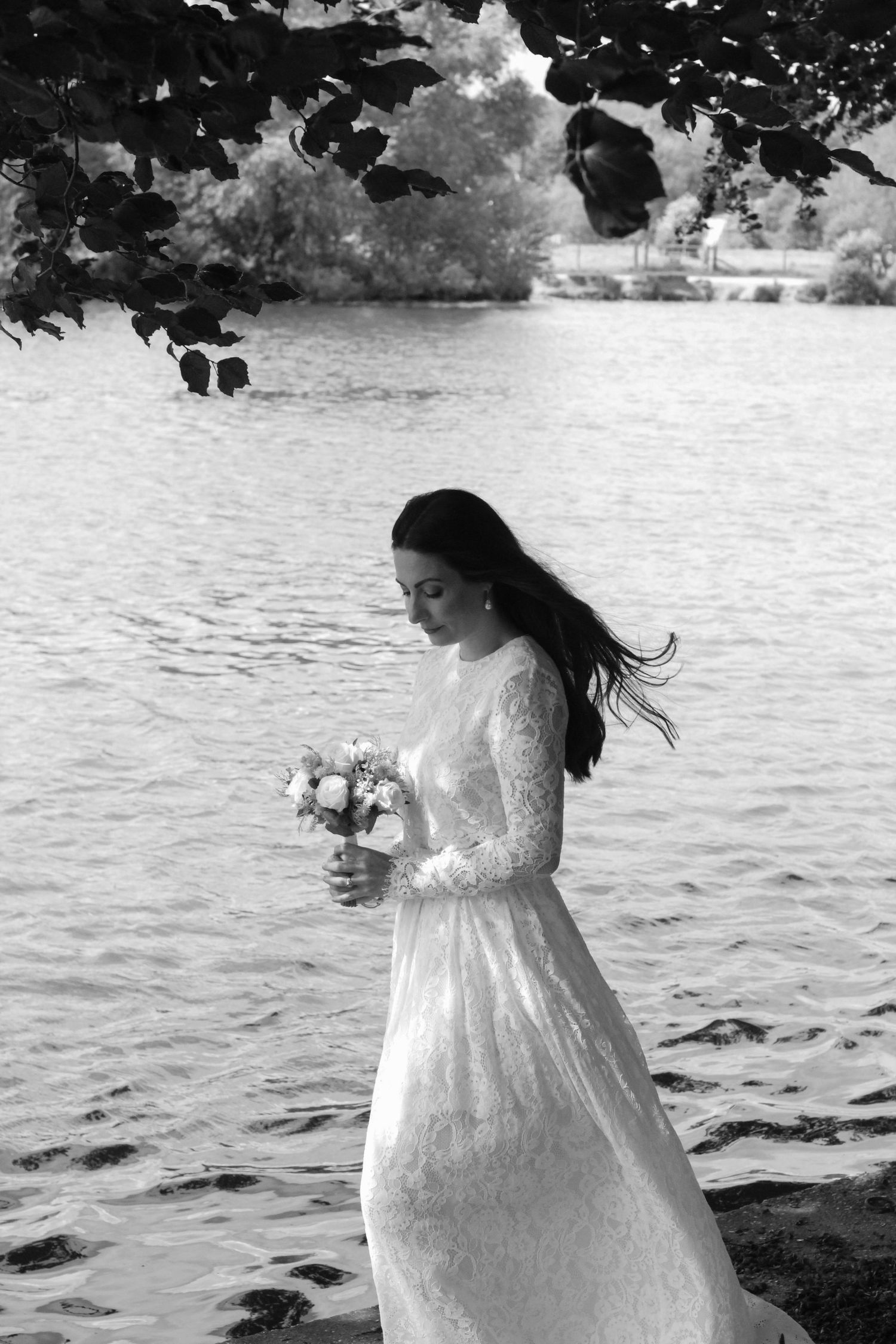 A bride in a lace wedding dress walks along a waterfront in black and white photography.