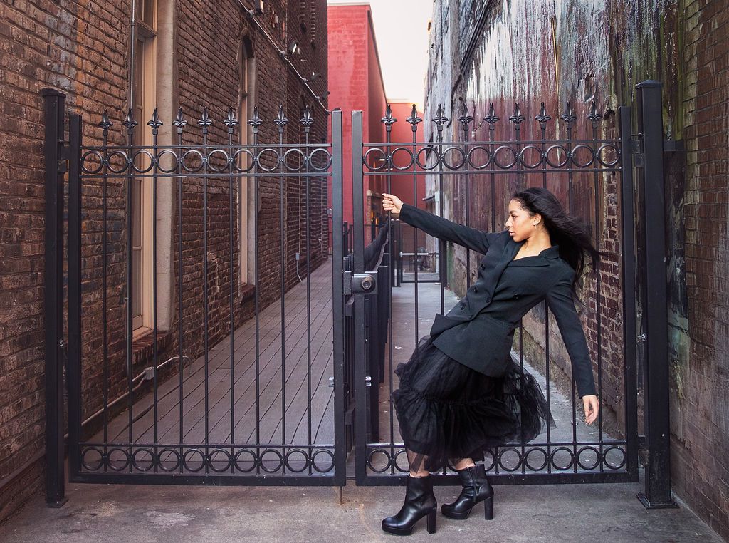 Teen girl dressed in all black with black hair leans back dramatically while holding black iron gate next to murals on Gay St. in Knoxville.