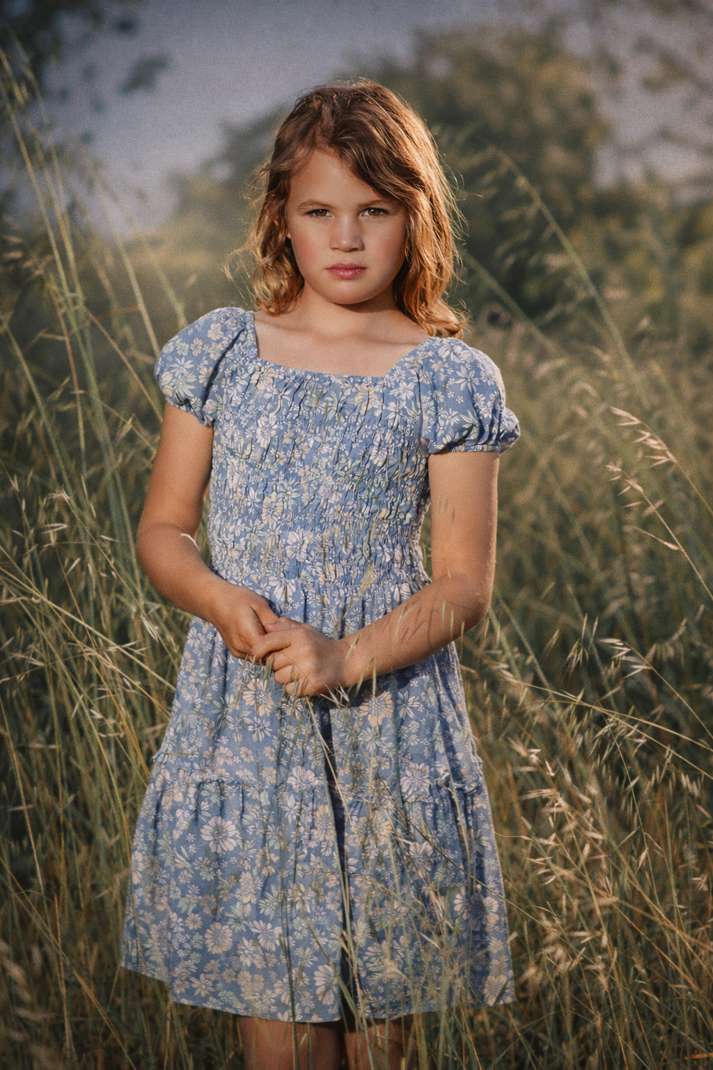 Young child in blue floral dress standing in tall summer grass during golden hour. Blue, Green. Brown Hair.