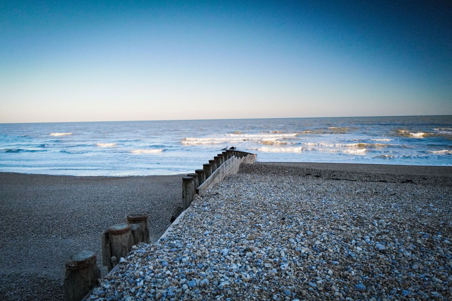 Misty blue coastal landscape with waves crashing on a pebble beach.