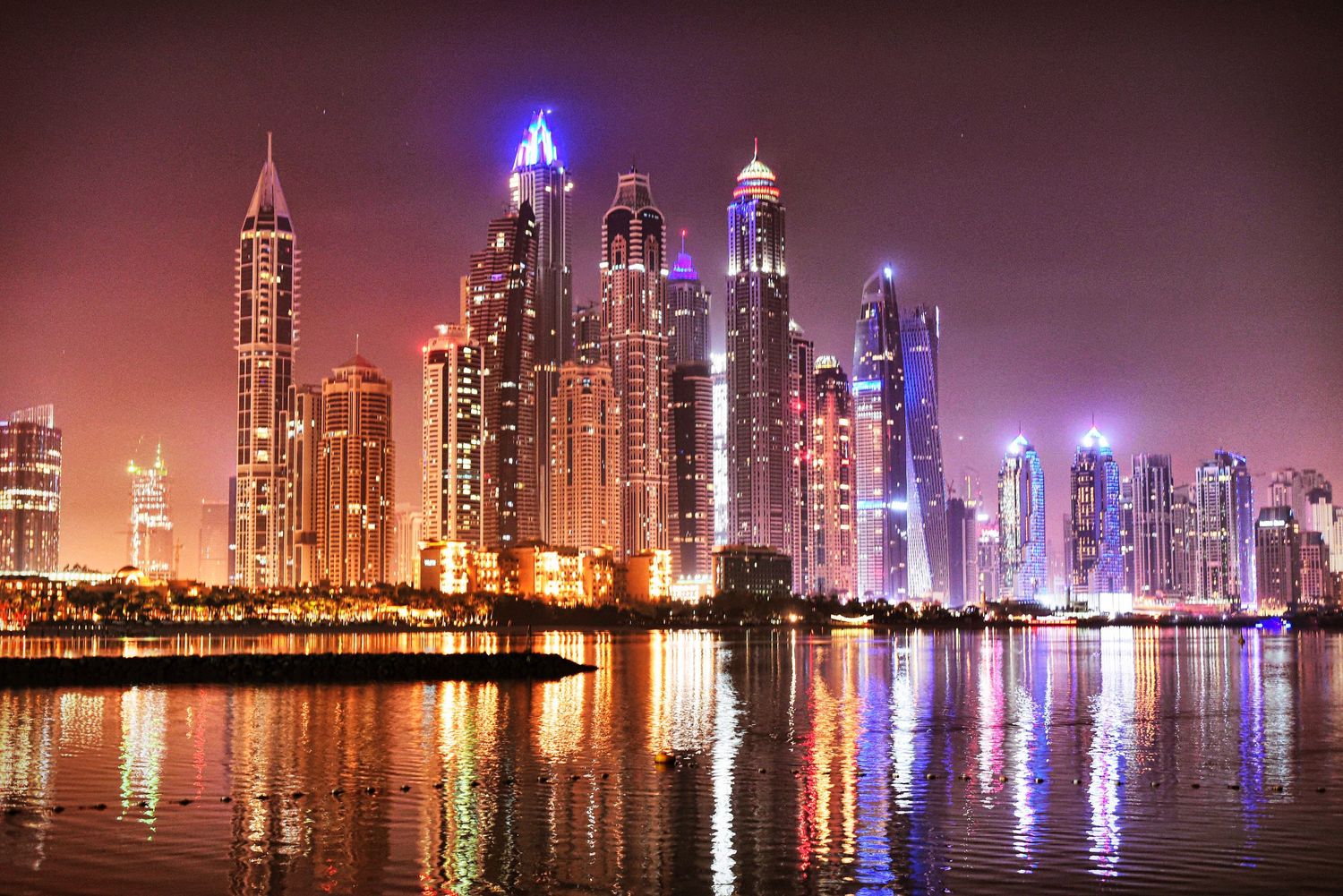 Illuminated Dubai city skyline at night reflecting in the water with colorful lights and modern skyscrapers.