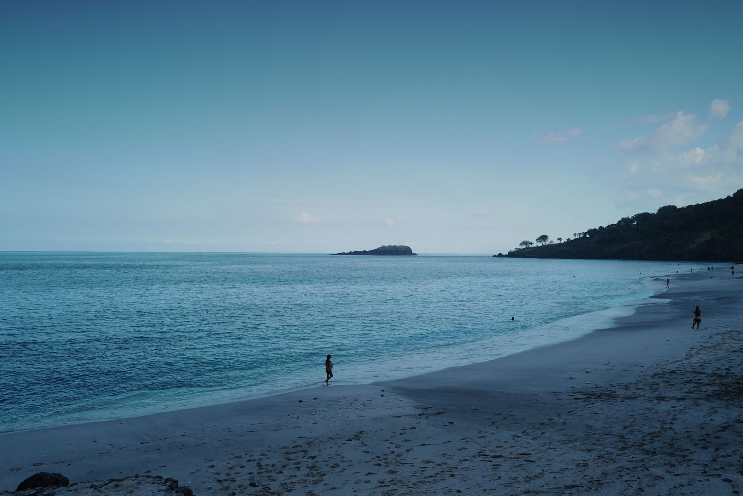 Serene blue coastal scene with lone figure walking along empty beach against distant headland.