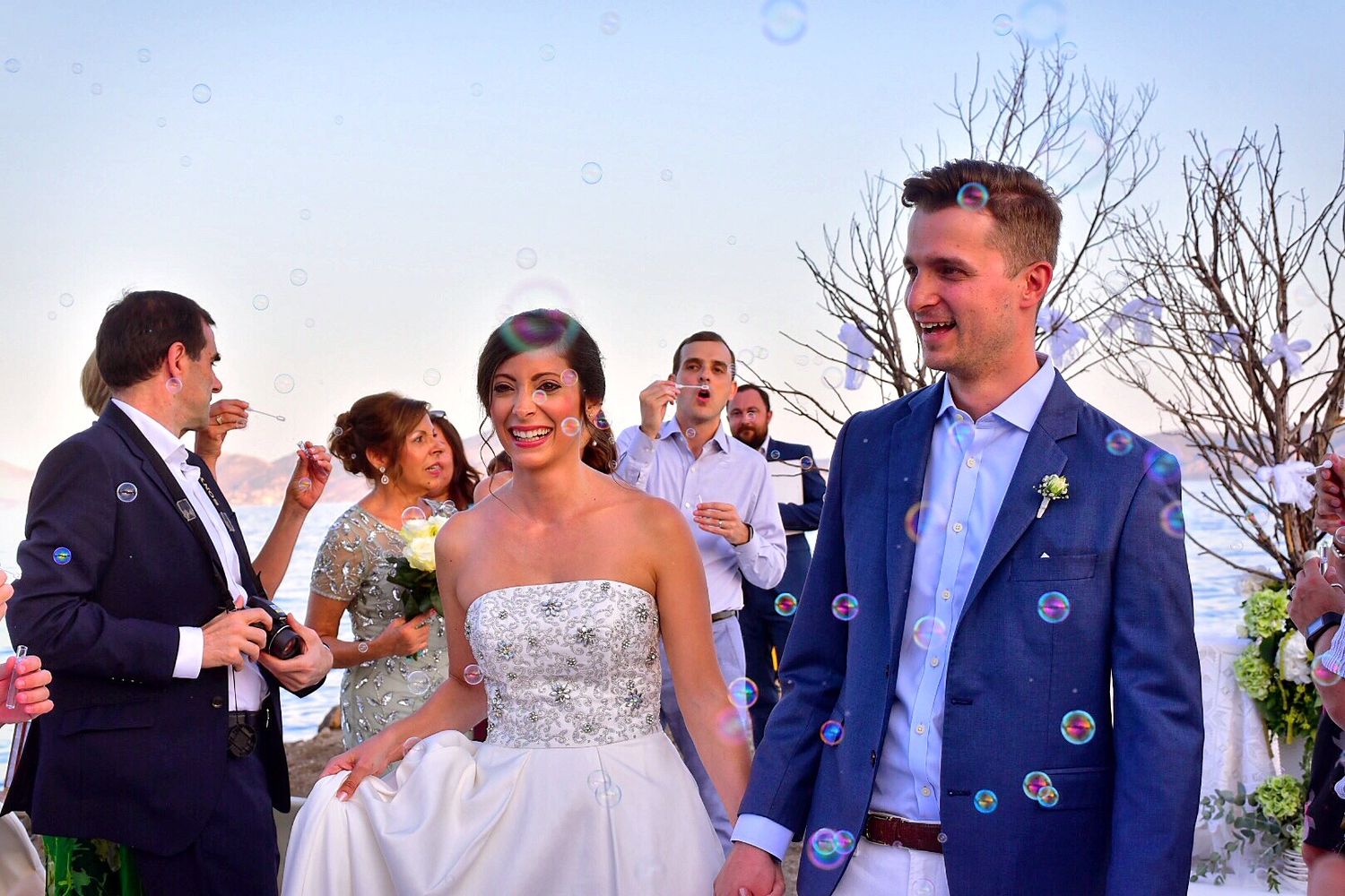Joyful wedding celebration with couple and guests under string lights with ocean backdrop.